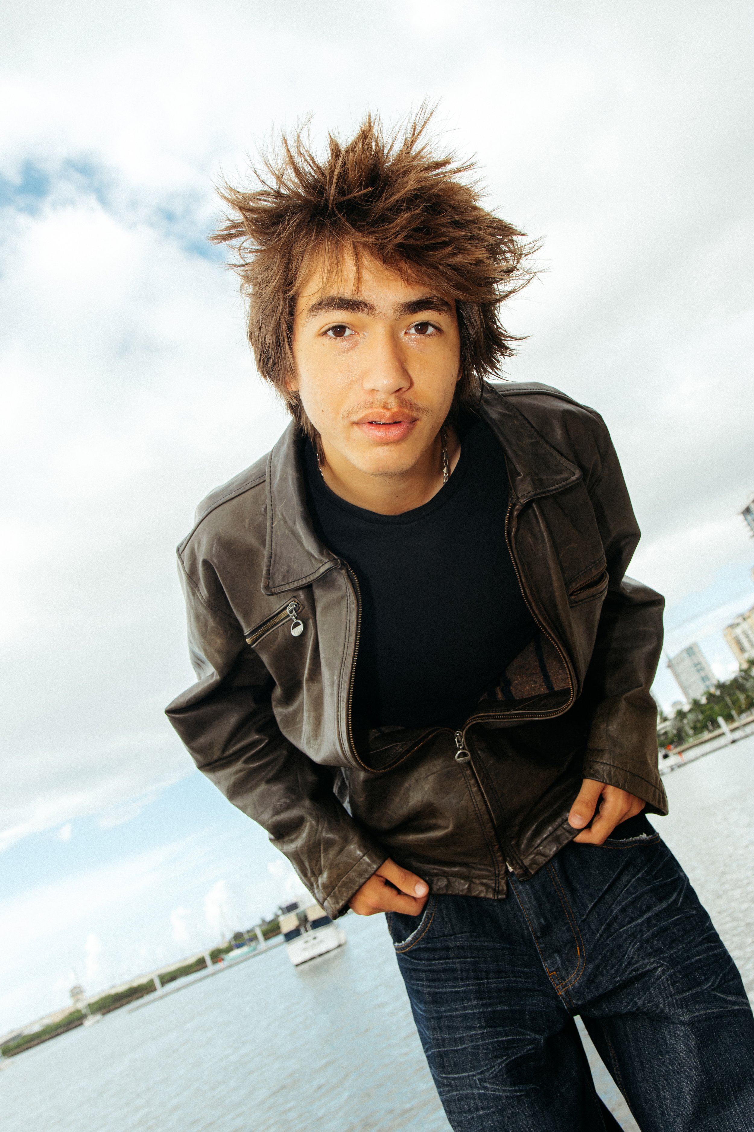 A young man with spiked brown hair wearing a black shirt, a brown leather jacket, and jeans, standing outdoors by water with boats and buildings in the background.