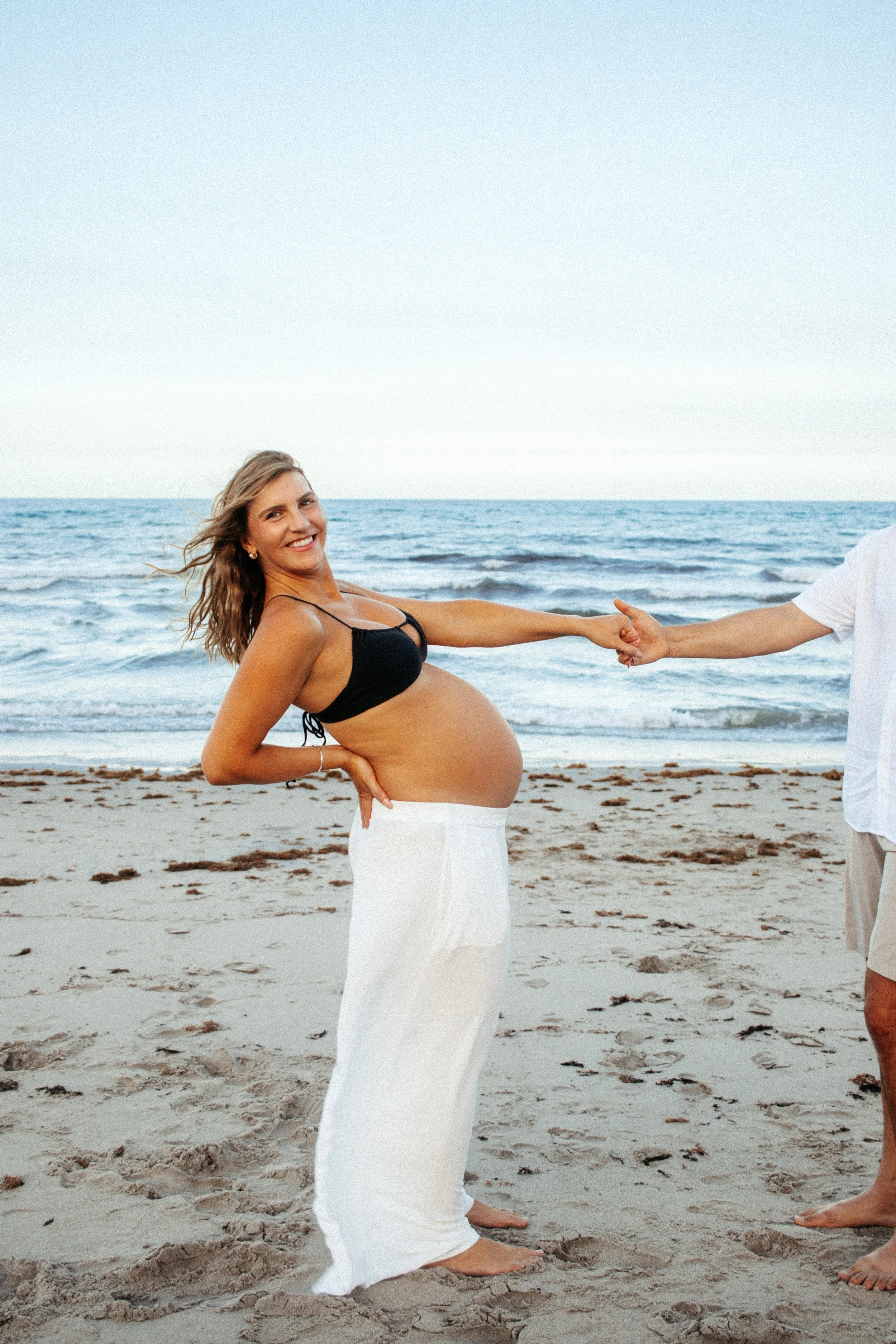 A pregnant woman smiling on the beach wearing a black bikini top and white skirt, holding hands with an unseen person, with the ocean and sand in the background during a Romantic Beach Couples Photography Session