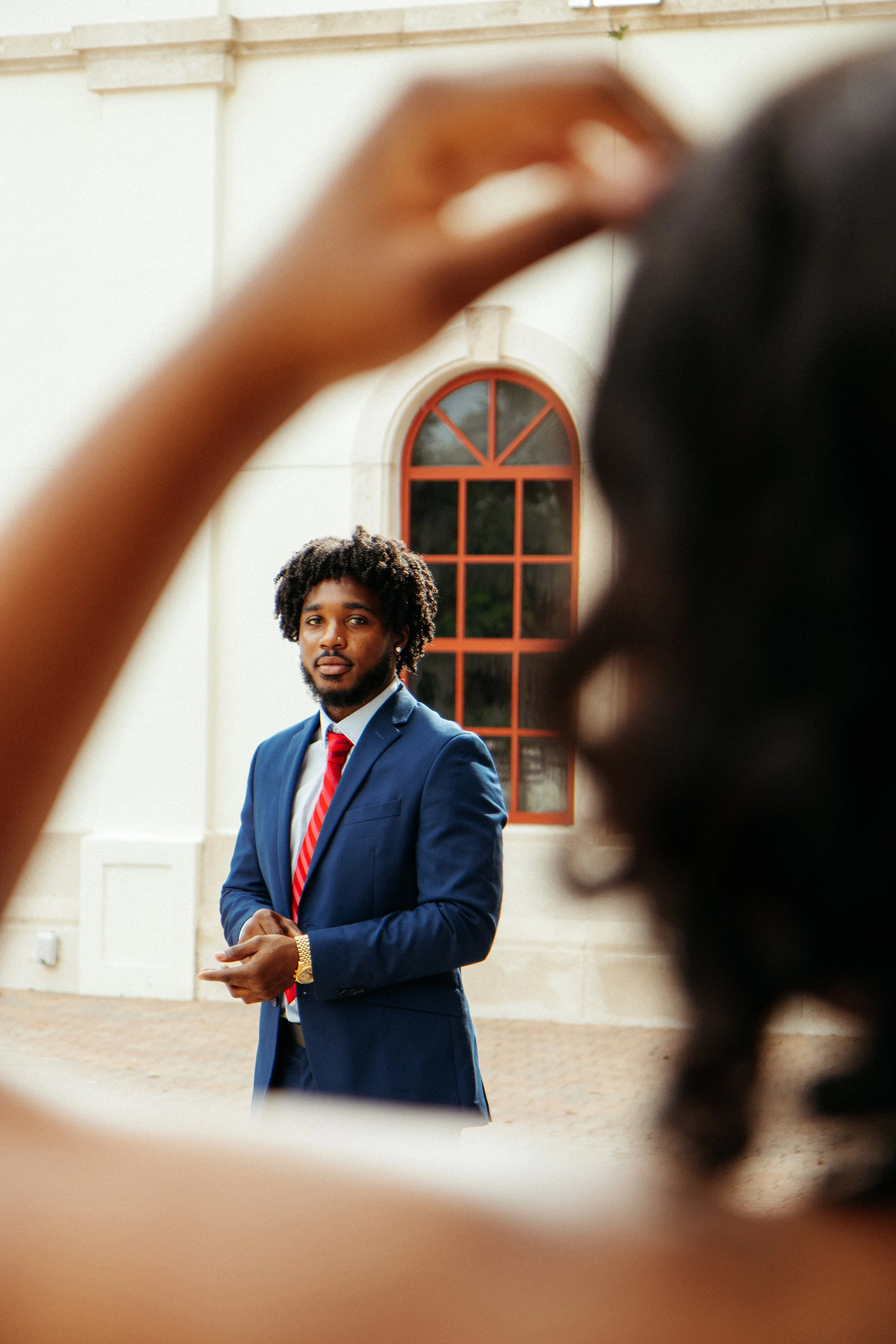 A man in a blue suit and red tie standing outside in front of a light-colored building with arched windows, seen through a blurred frame and a person with dark skin and curly hair in the foreground.