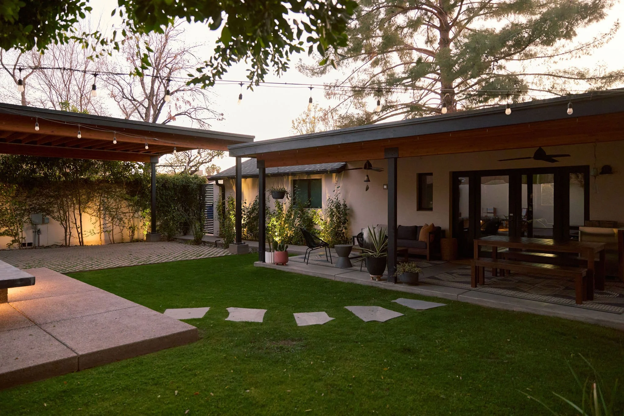 A backyard patio with outdoor seating, potted plants, string lights overhead, and a lawn with stepping stones, adjacent to a house with a covered porch and large glass doors.