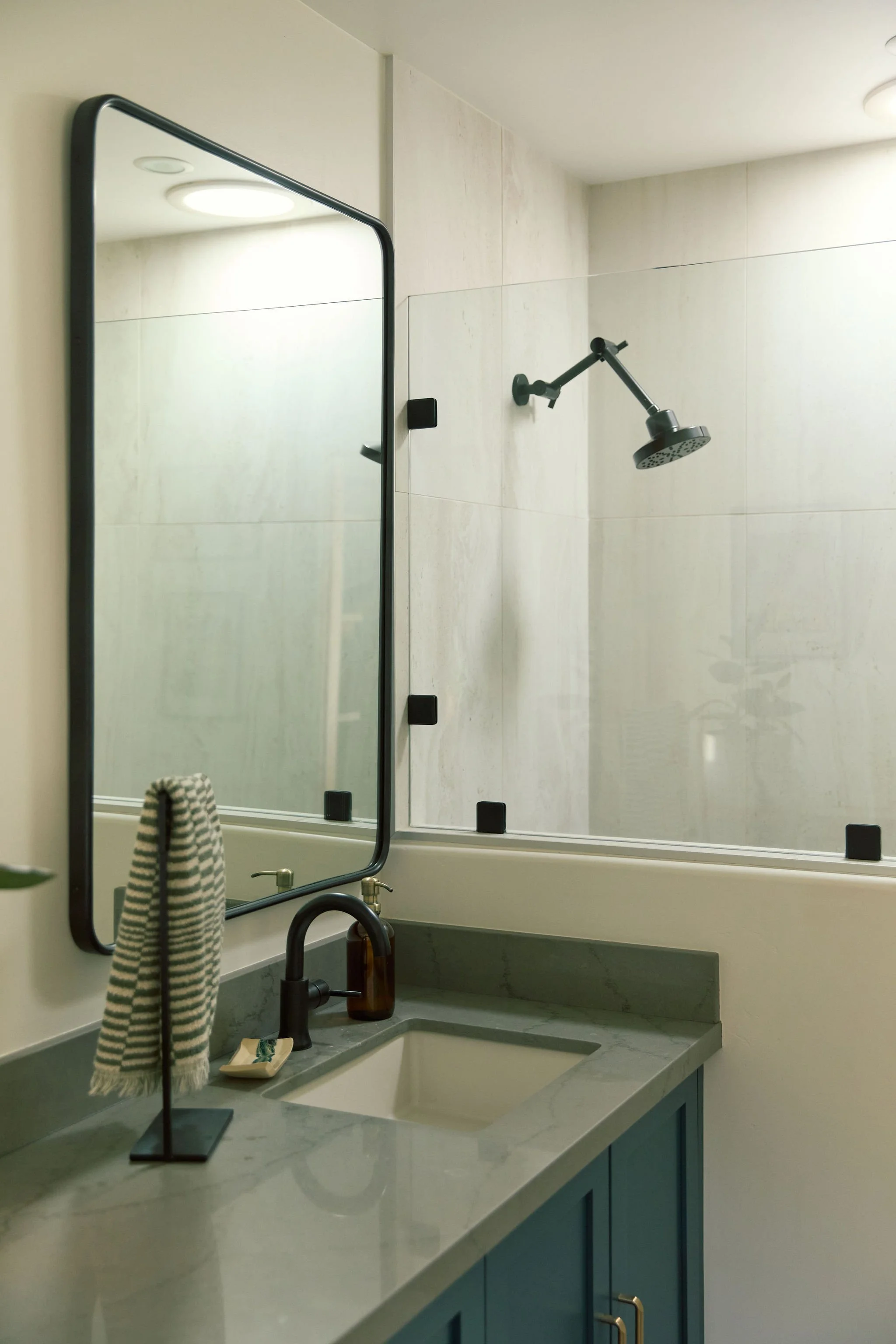 Modern bathroom with a mirror, a green countertop with a beige sink, black faucet, soap dispenser, striped towel on a stand, and a glass partition with a black frame. Showerhead on the wall.