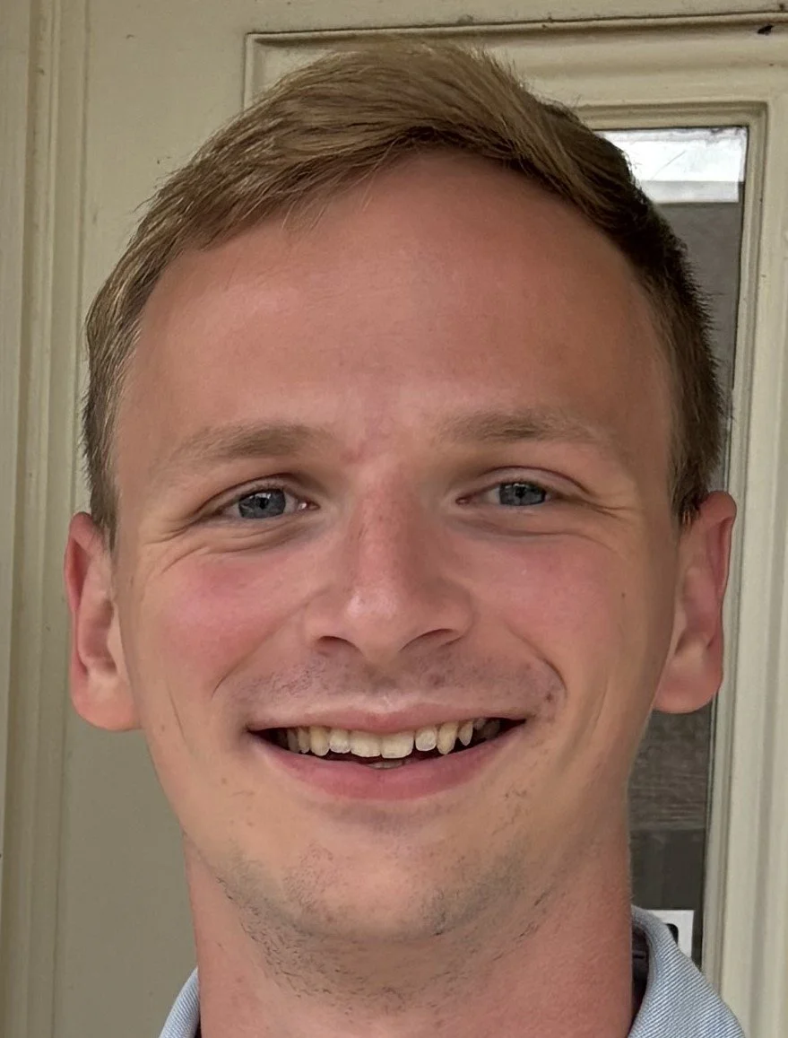 Close-up of a young man smiling, showing his teeth, with light skin, short light brown hair, blue eyes, and a background of a door.