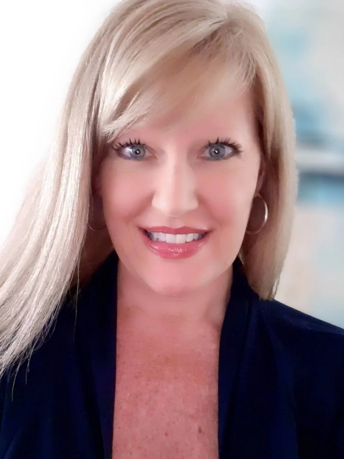 A woman with long blonde hair, blue eyes, and light makeup, smiling at the camera. She is wearing hoop earrings and a dark top.