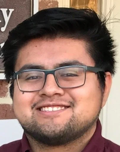 A young man with black hair and glasses smiling in front of a brick wall.