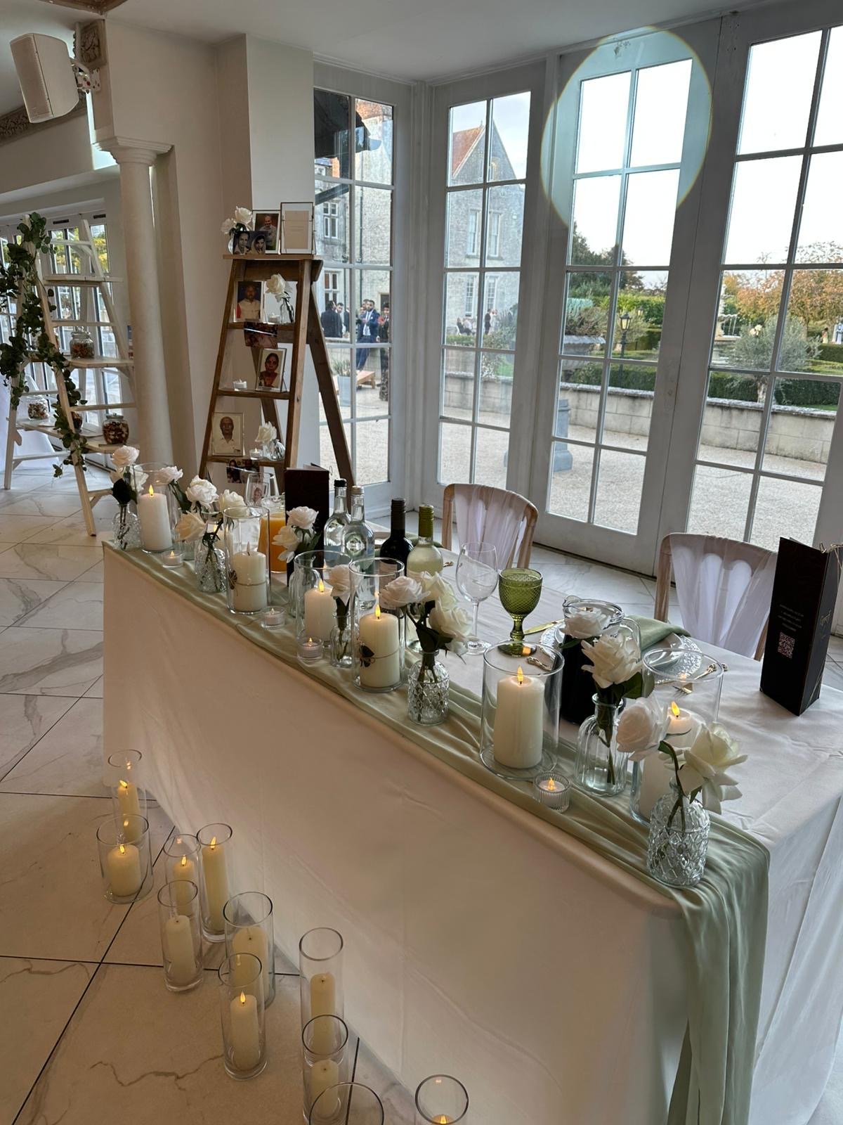 Inside a bright, elegant room decorated for a wedding reception or celebration with large windows, a table with white flowers, candles, and bottles, and photo frames on a wooden ladder.