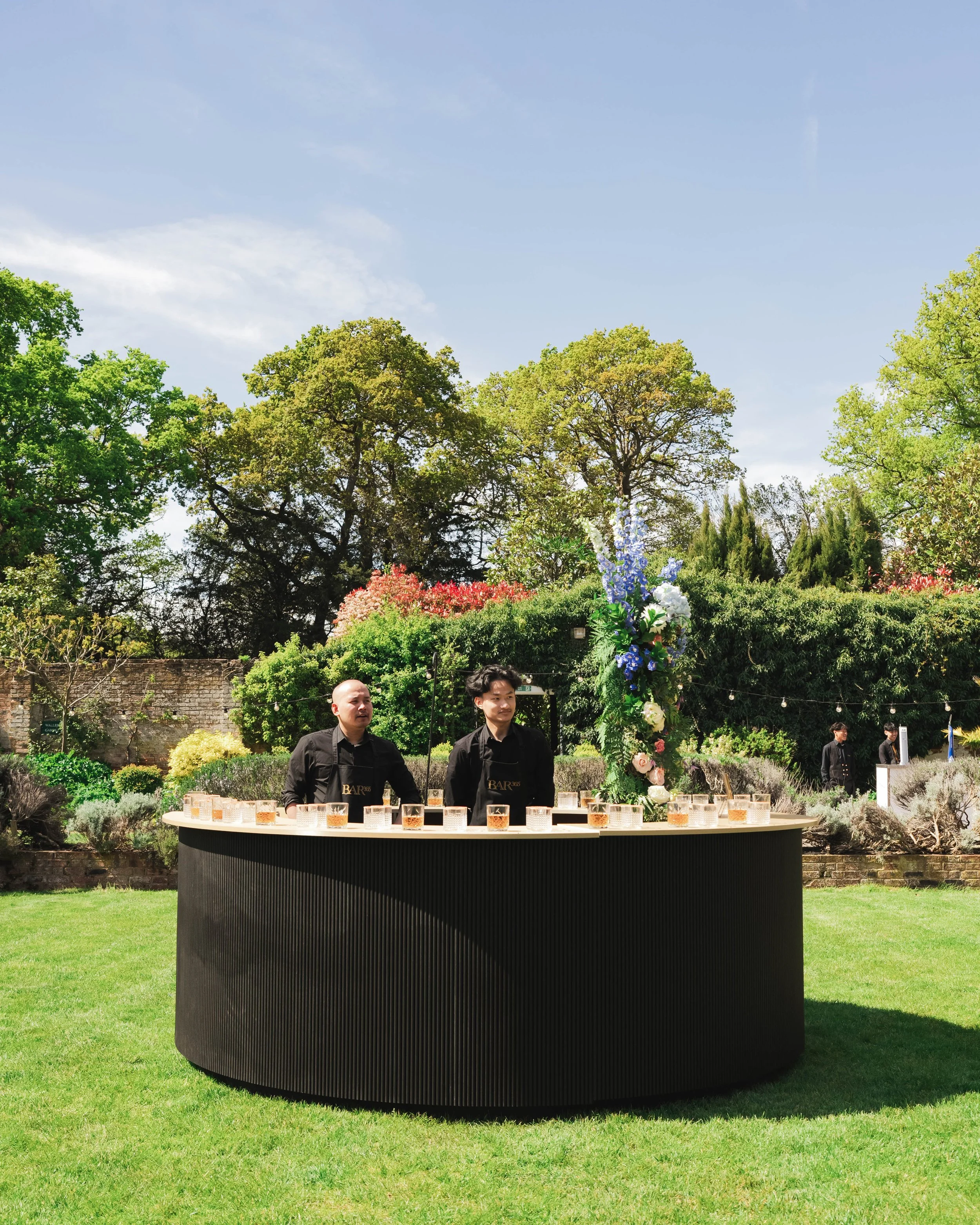 Outdoor bar setup with two bartenders dressed in black, serving drinks at a curved bar counter decorated with a large floral arrangement, amid a lush garden with trees and a blue sky. Hampshire & Fleet Indian & Nepalese GuruJi Guru-Ji restaurant