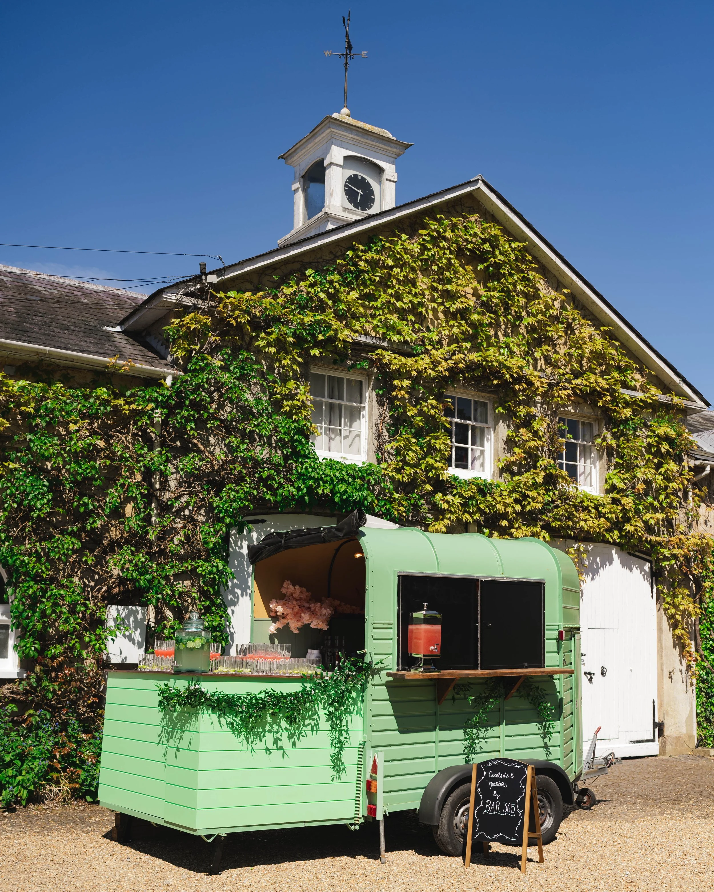 Green mobile bar trailer in front of a ivy-covered building with a clock tower, under a clear blue sky. Hampshire & Fleet Indian & Nepalese GuruJi Guru-Ji restaurant