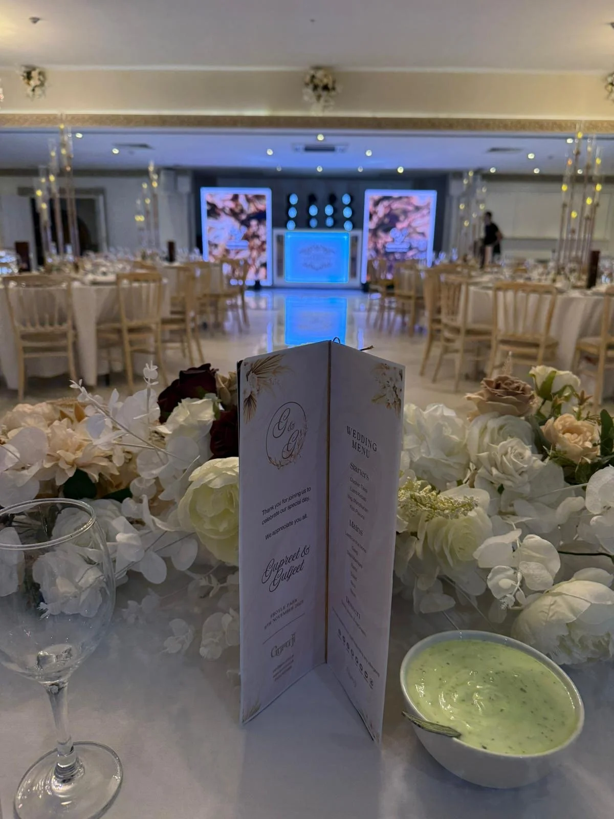 Wedding reception hall with decorated tables, floral arrangements, a wedding menu, water glass, and green dip in bowl in the foreground, with a stage and screens in the background.