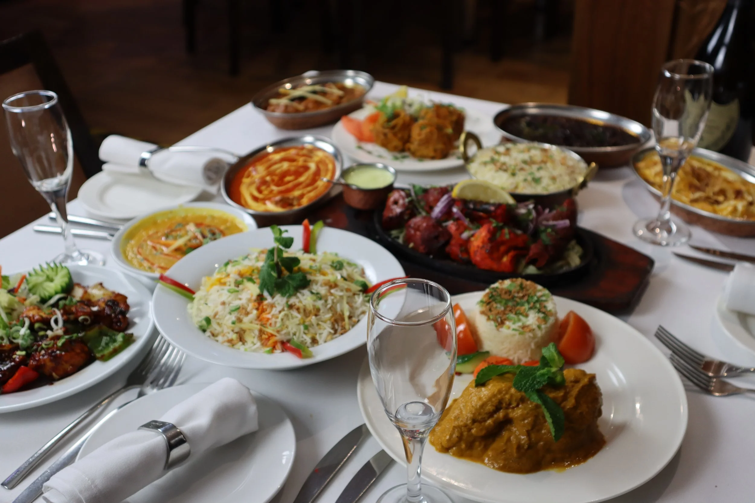 An Indian feast with various dishes on a white tablecloth, including rice, curry, grilled meats, naan, and assorted side dishes, with empty glasses and cutlery.