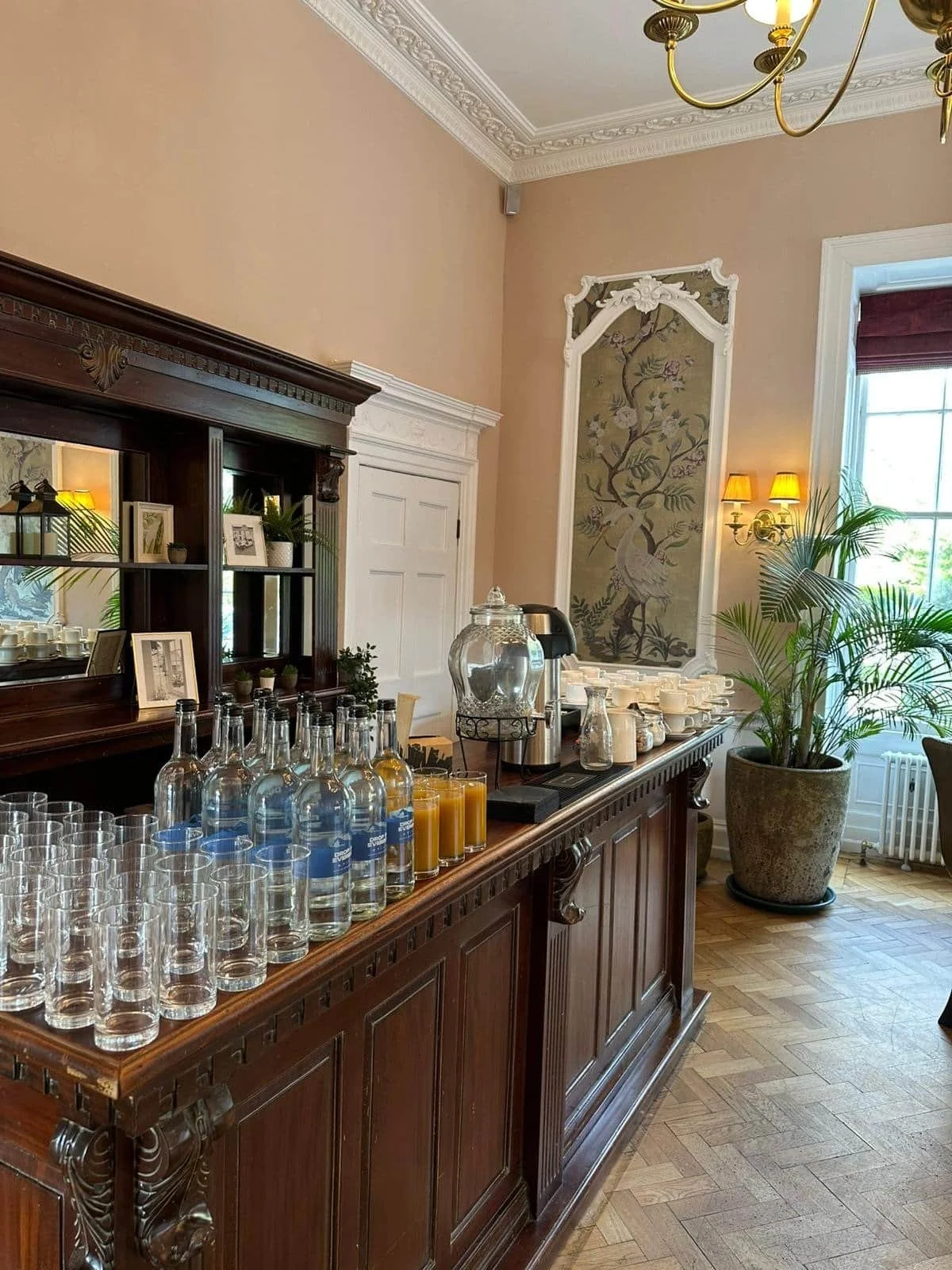 A wooden sideboard with glass bottles of water and orange juice, a large glass beverage dispenser, cups, and coffee pots in a well-lit room with a large window, potted plant, decorative wall art, and a chandelier.
