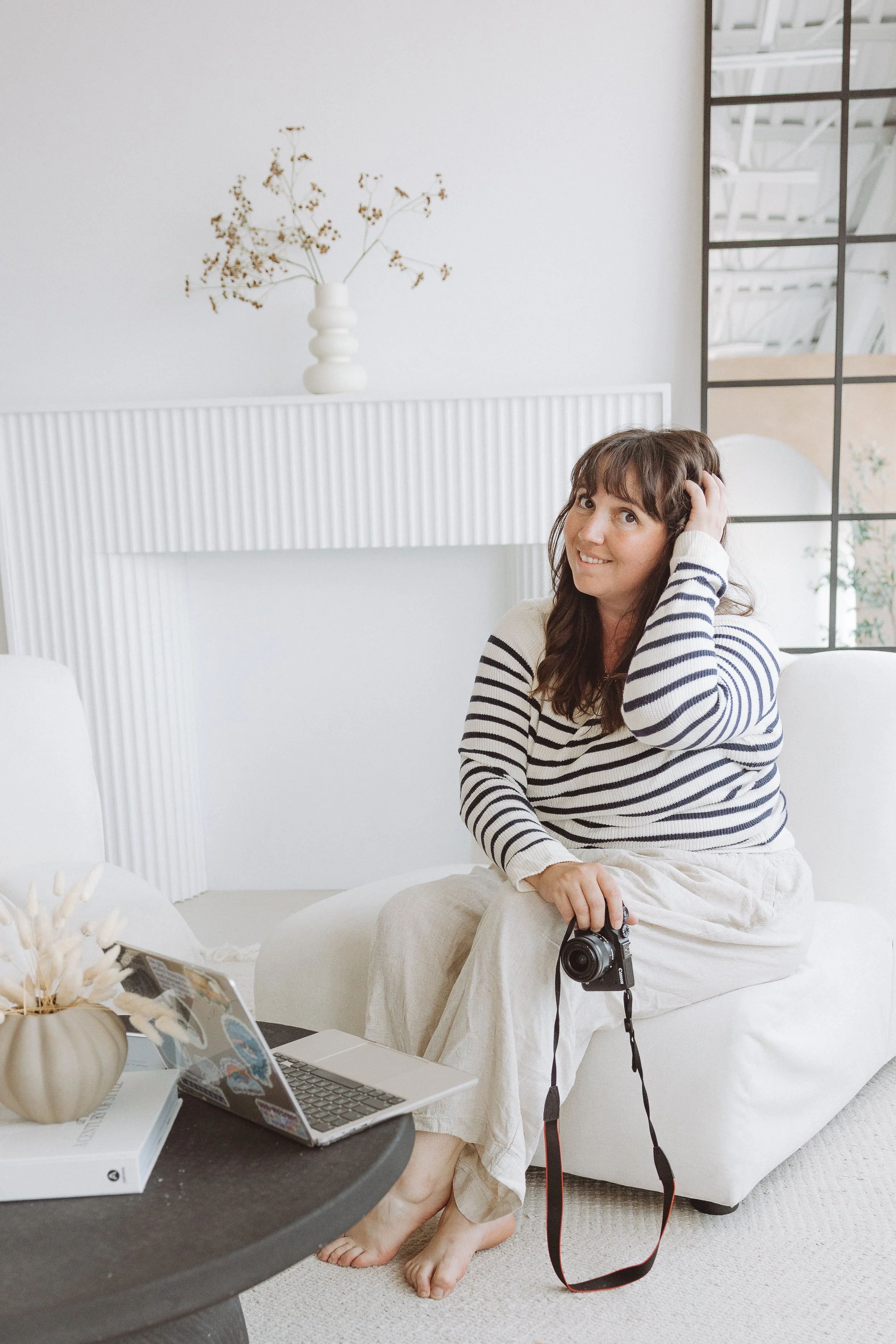 A woman with brown hair and bangs sitting on a white sofa in a bright, modern room. She is smiling, wearing a striped sweater and beige pants, holding a camera in her lap, with a laptop and a book on a black coffee table in front of her. There's a vase with dried flowers on the table, and a decorative white vase with flowers on the white mantel behind her. A large mirror is on the wall to her right.