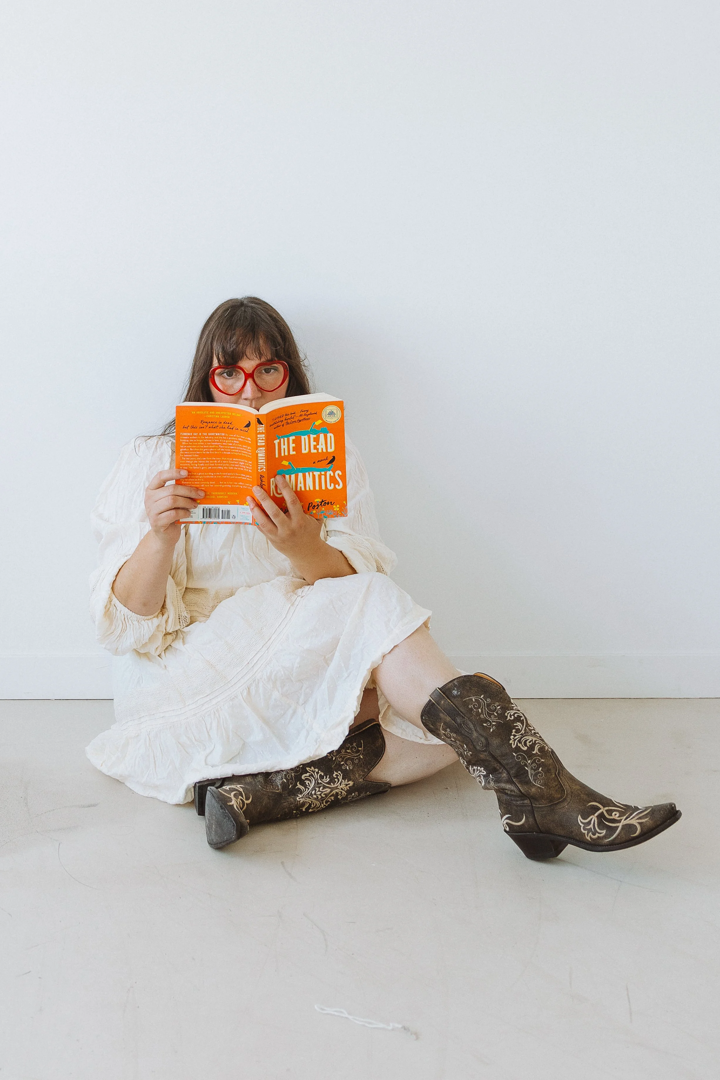 Woman with red glasses sitting on the floor reading a book titled 'The Dead Romantics' while wearing cowboy boots and a white dress in Toronto.