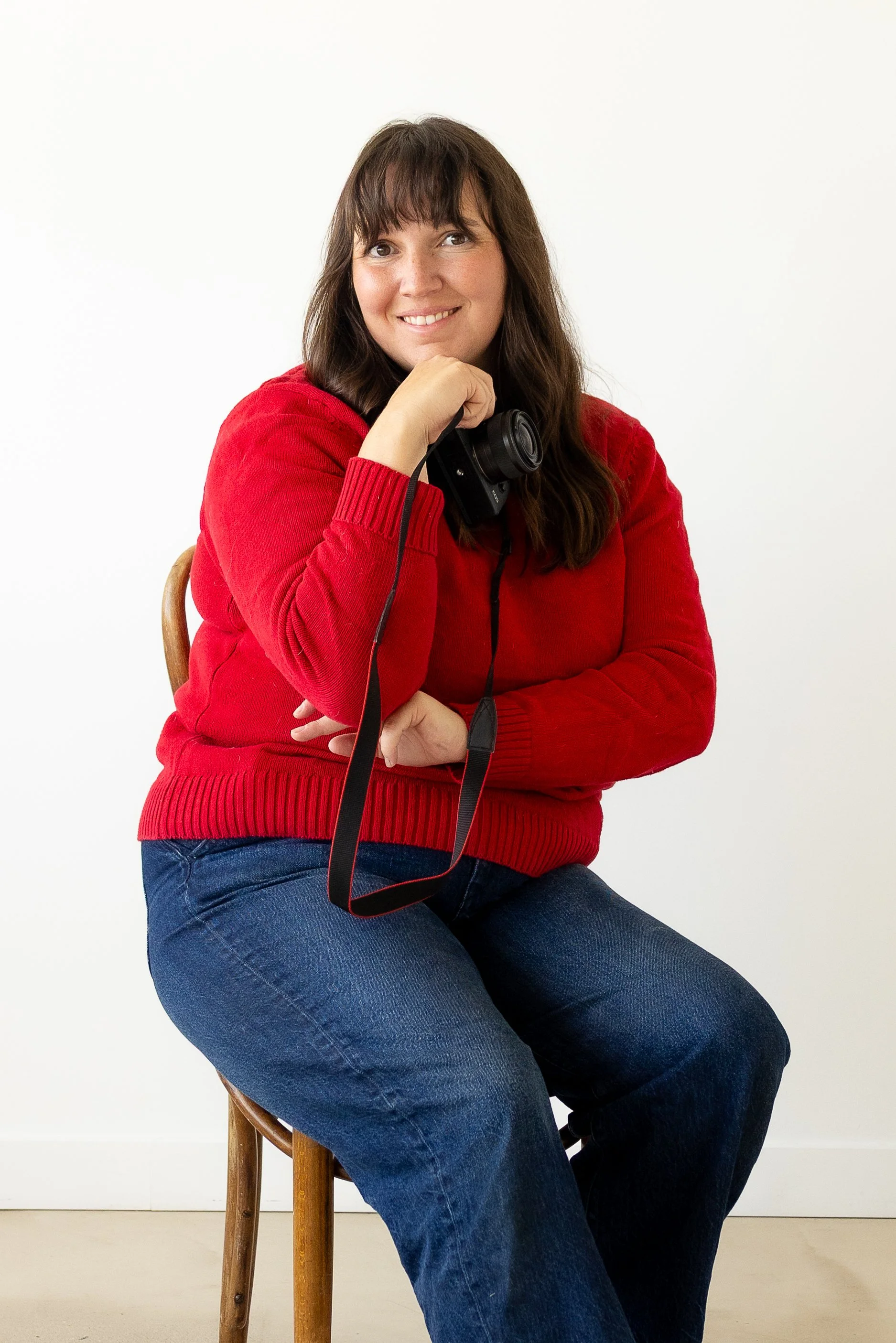 In a Toronto studio, a woman with long brown hair and a red sweater sitting on a wooden chair, holding a camera, against a plain white background.