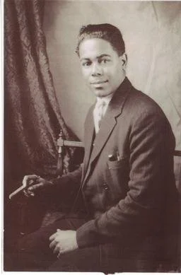 Black and white portrait of Denver Ferguson in a suit sitting with a cigarette in hand, in front of a curtain backdrop.
