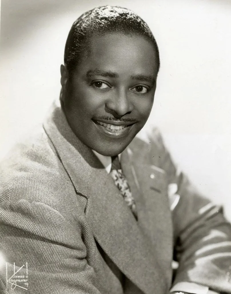 Black and white portrait of a smiling man wearing a suit jacket, dress shirt, and tie, with short hair and a mustache.
