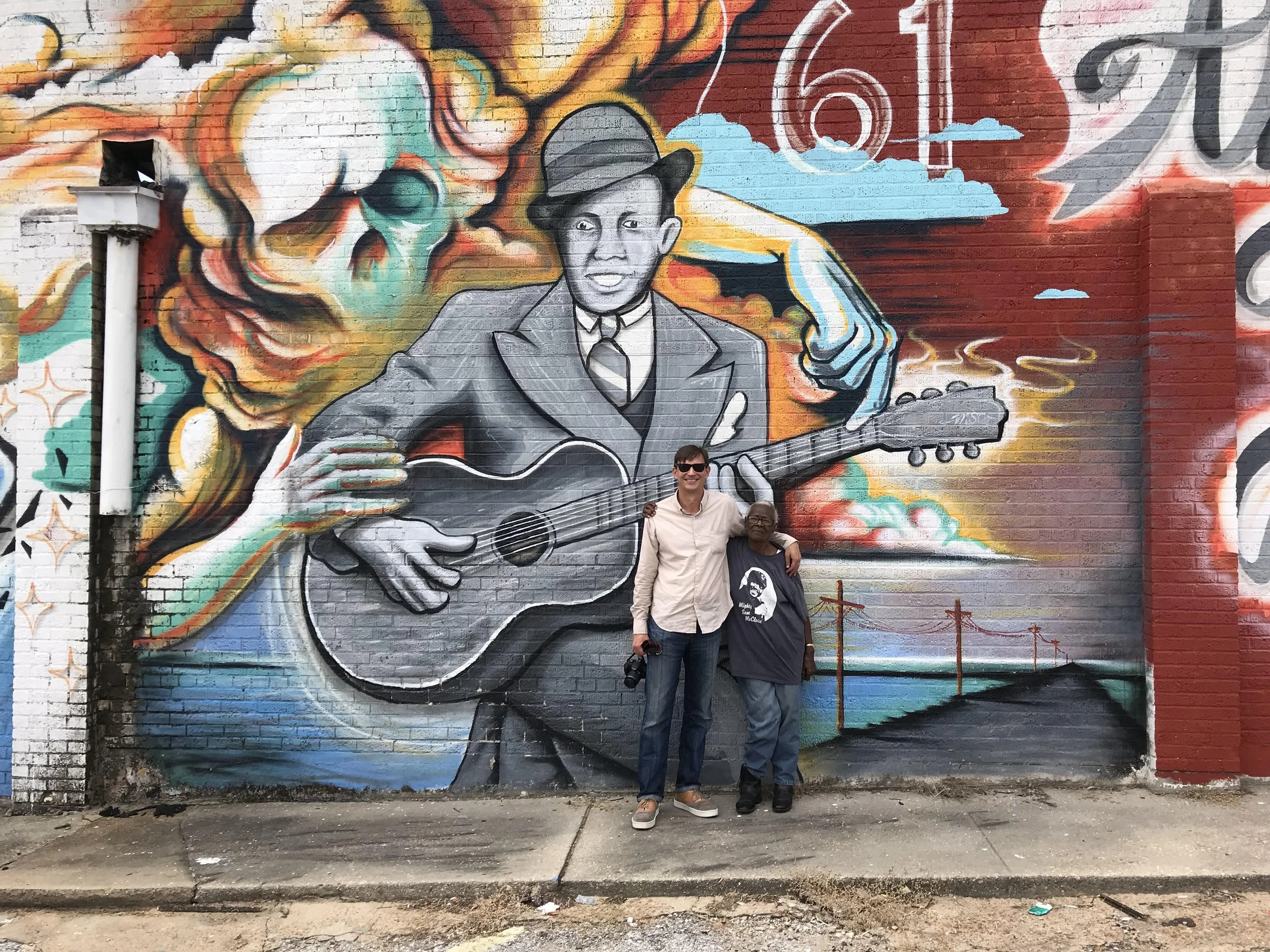 A mural depicting bluesman Robert Johnson with a hat playing a guitar, with colorful abstract and cloud-like background elements. Preston Lauterbach and Annye Anderson stand in front, with their arms around each other