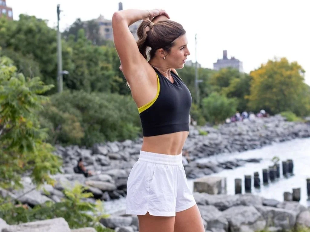 A young woman in athletic clothing stretching outdoors near a rocky shoreline with trees and water in the background.
