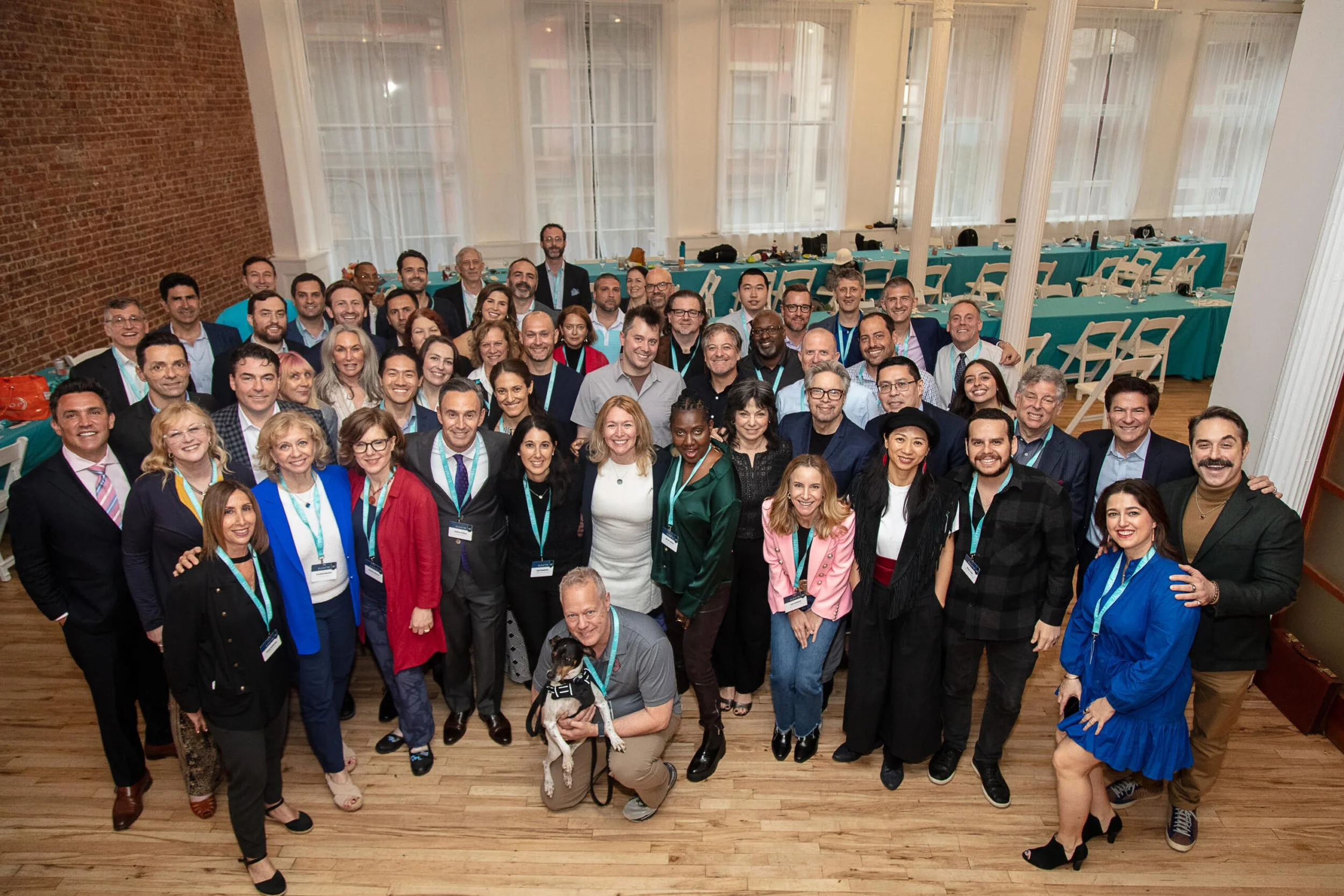 A large group of people dressed in business attire gathered for a group photo in a spacious room with large windows, a brick wall, and teal tablecloths in the background. A man crouching in the front holds a dog.