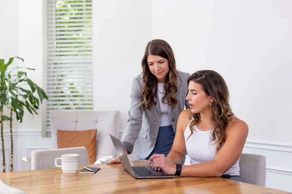 Two women are talking at a wooden table in an office. The woman on the right is seated, working on a laptop, while the woman on the left is standing and pointing at the screen. There is a white coffee mug and a smartphone on the table, and a large window with blinds and a plant in the background.