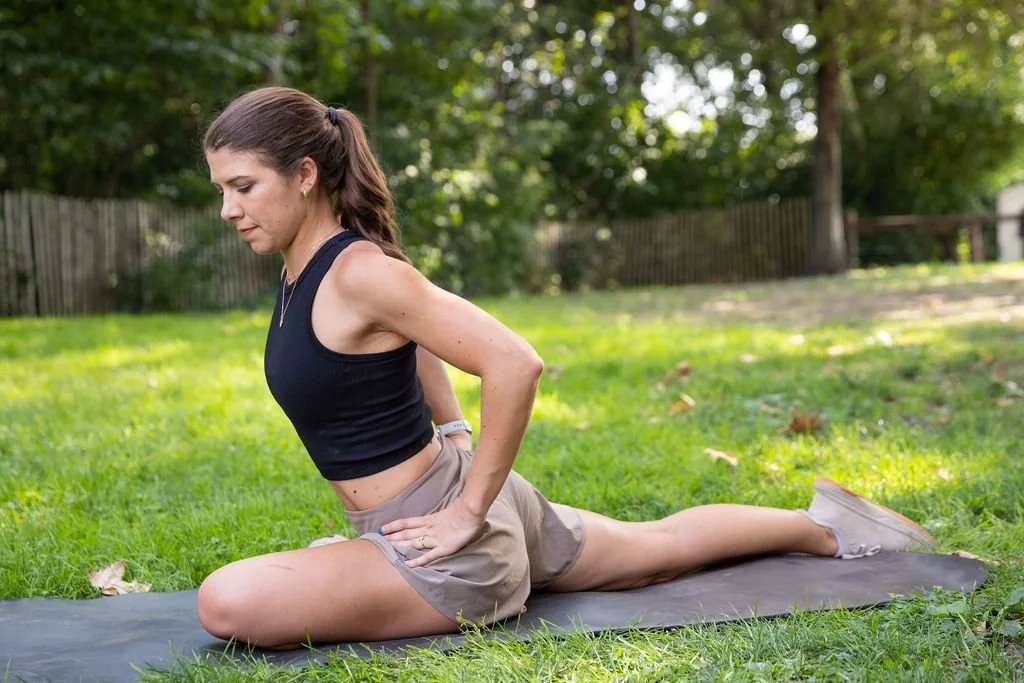 A woman practicing yoga outdoors on a grassy field with trees and a wooden fence in the background. She is in a lunge position on a yoga mat, wearing a black tank top and beige shorts.
