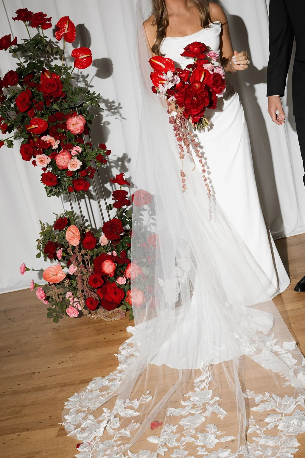 Bride in a white wedding dress holding a bouquet of red and pink flowers, standing next to a floral arrangement of red, pink, and peach blooms.