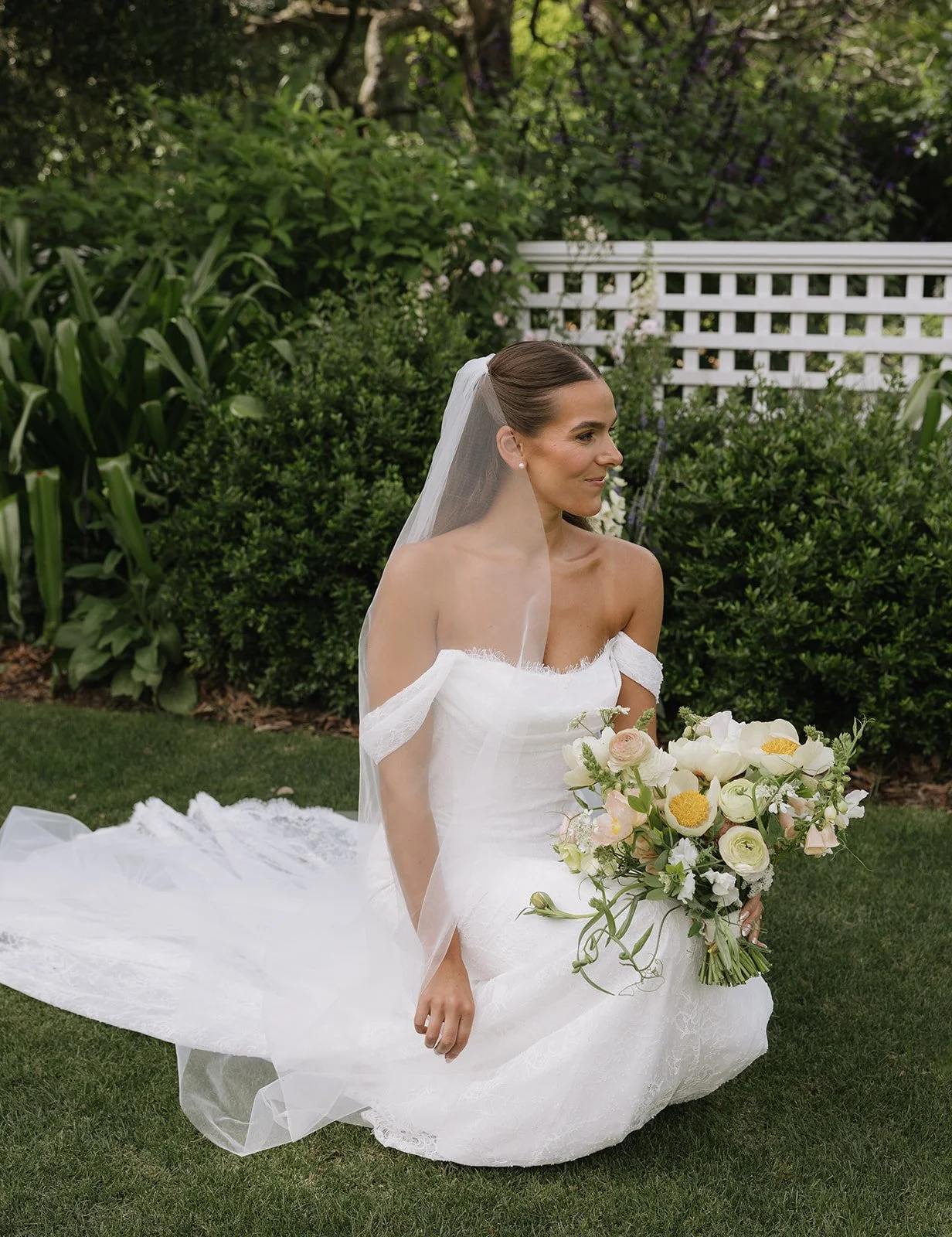 A bride sitting on the grass in a garden, wearing a white off-the-shoulder wedding gown with a long veil, holding a bouquet of white, cream, and yellow flowers.