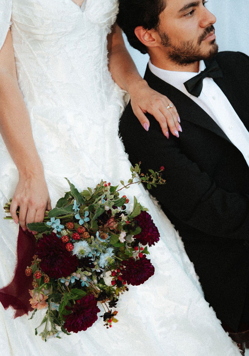 A bride in a white wedding dress holds a colorful bouquet of flowers, standing next to a groom in a black tuxedo with a bow tie, with her hand resting on his shoulder.