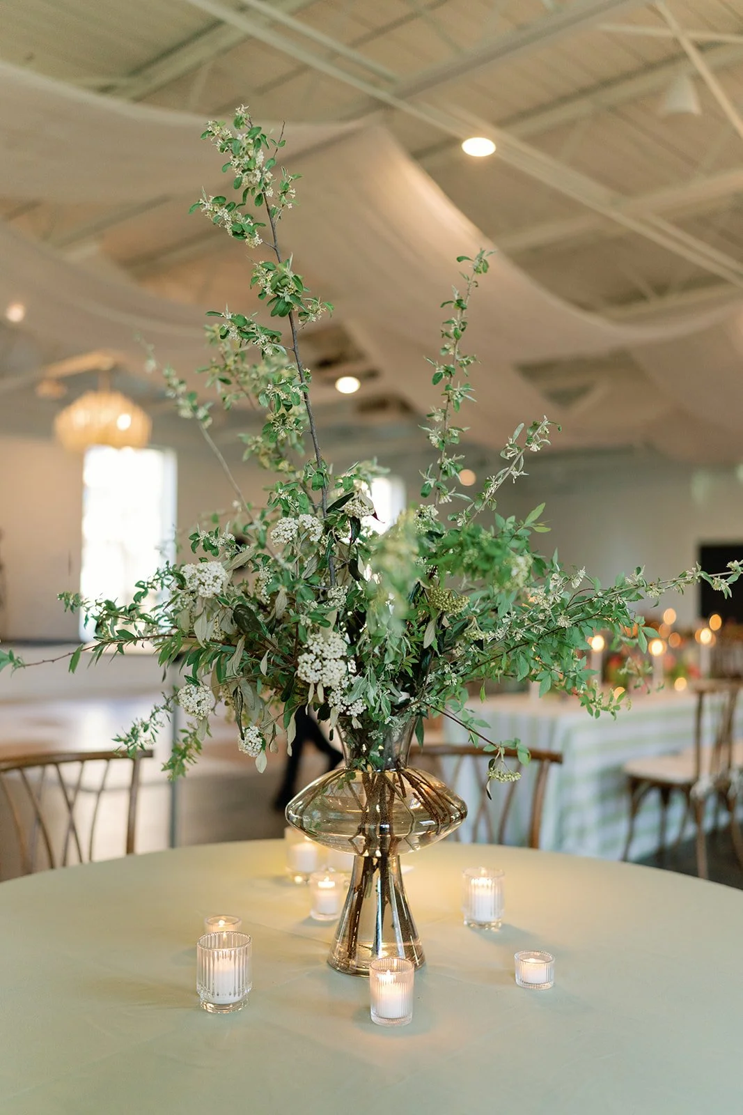 A floral centerpiece with green leaves and white flowers in a tall glass vase on a table decorated with small candleholders, set in a decorated indoor space.