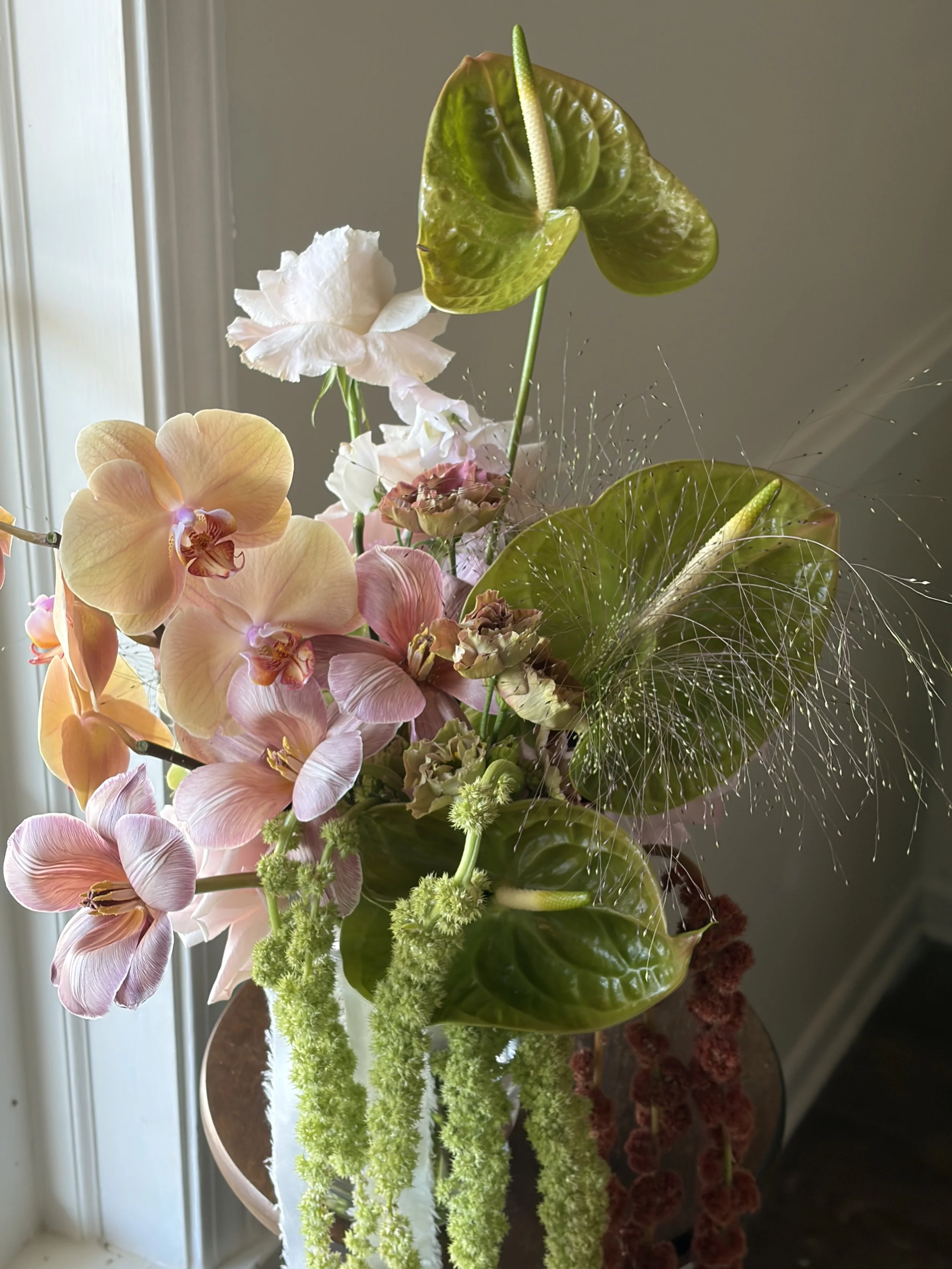 A floral arrangement with pink, white, and peach orchids, green anthuriums, and red-berry clusters in a vase near a window.