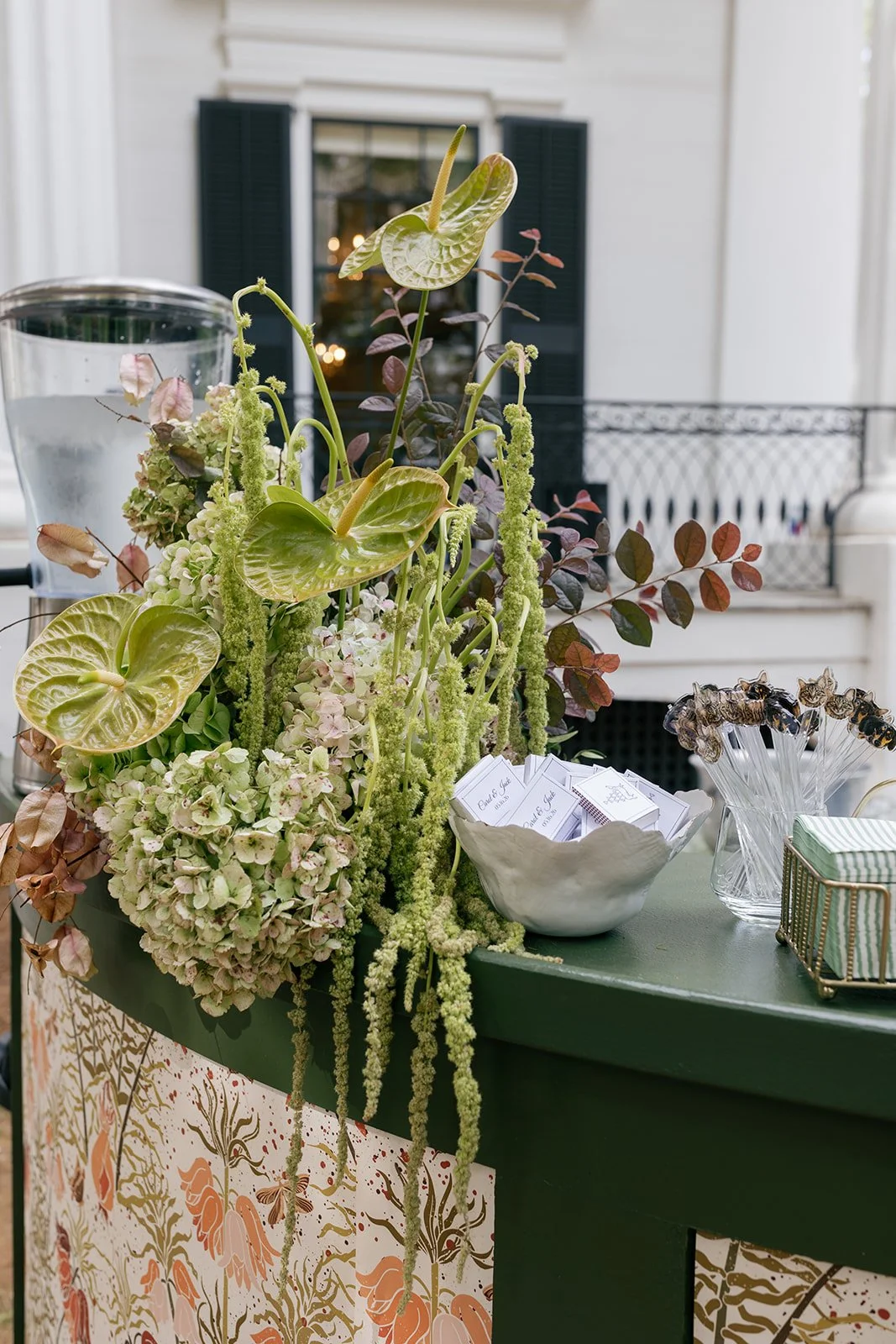 A floral arrangement with green and white flowers and plants on a green table, with a mason jar, cards, and decorative items nearby, set on a porch or patio with window and black shutters in the background.