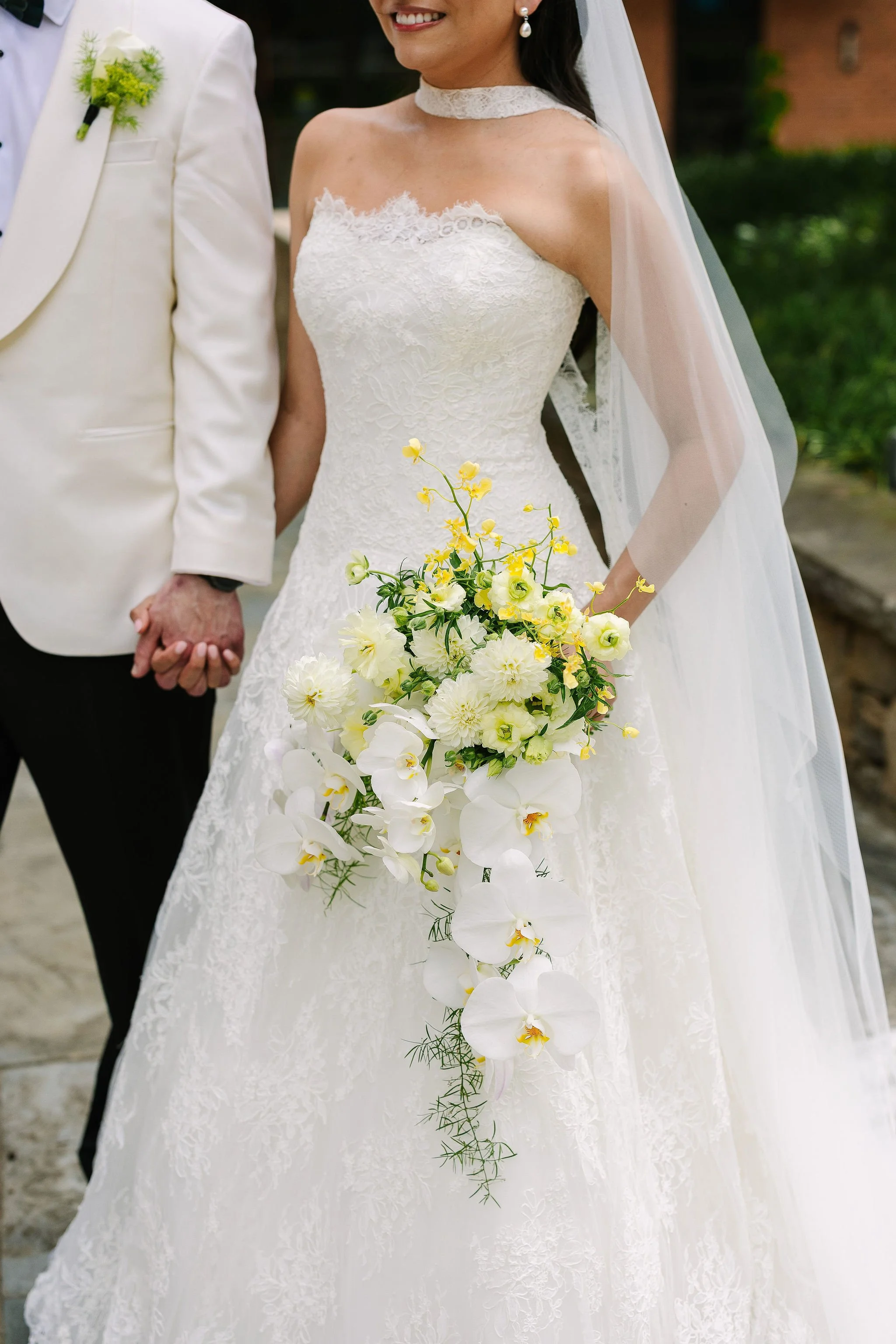 A bride in a white lace wedding dress holding a cascading bouquet of white and yellow flowers, standing next to a groom in a cream tuxedo with a white shirt and a green boutonniere.