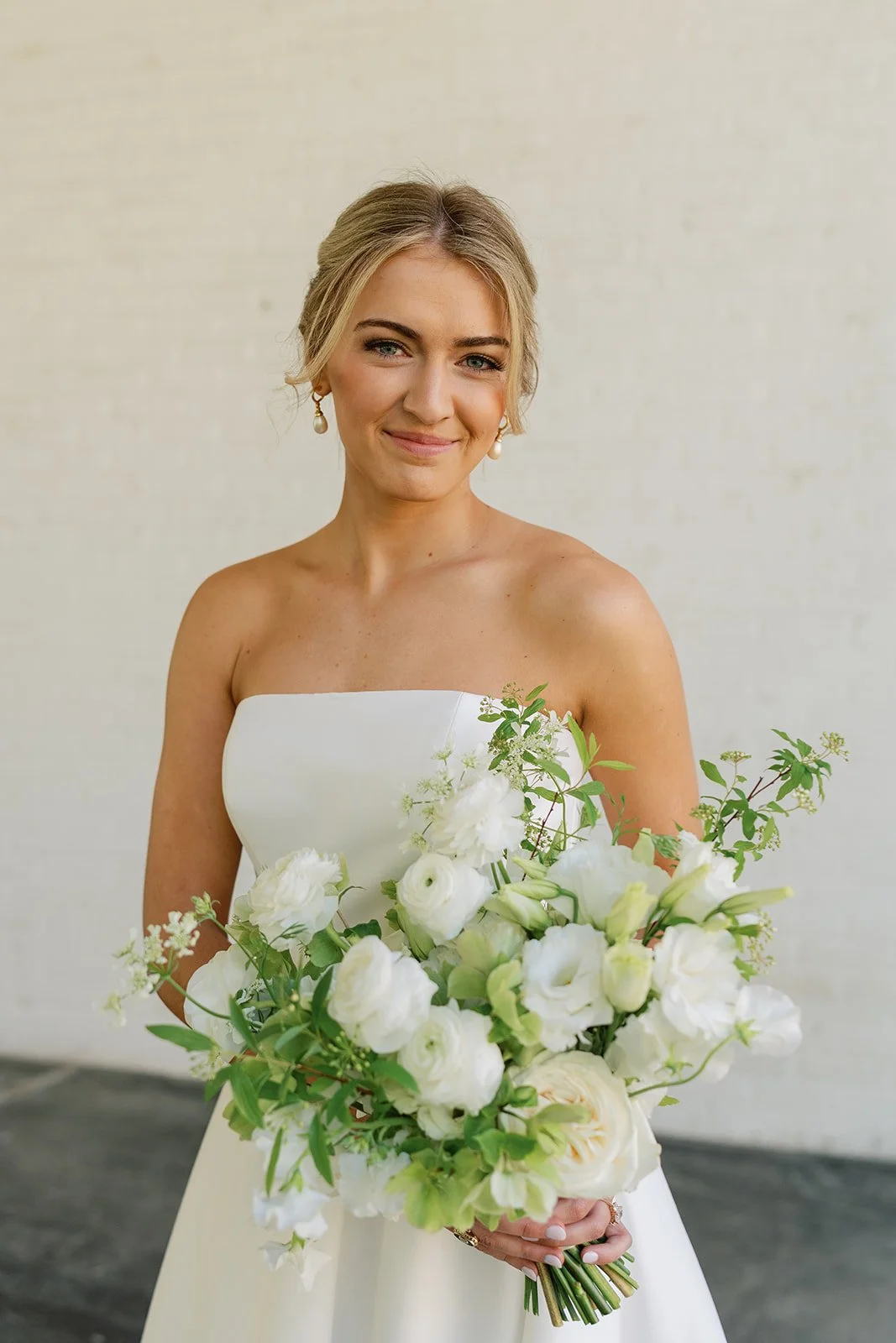 Smiling bride holding a bouquet of white flowers and greenery, dressed in a strapless white wedding gown, with her hair styled in an elegant updo and wearing pearl earrings.