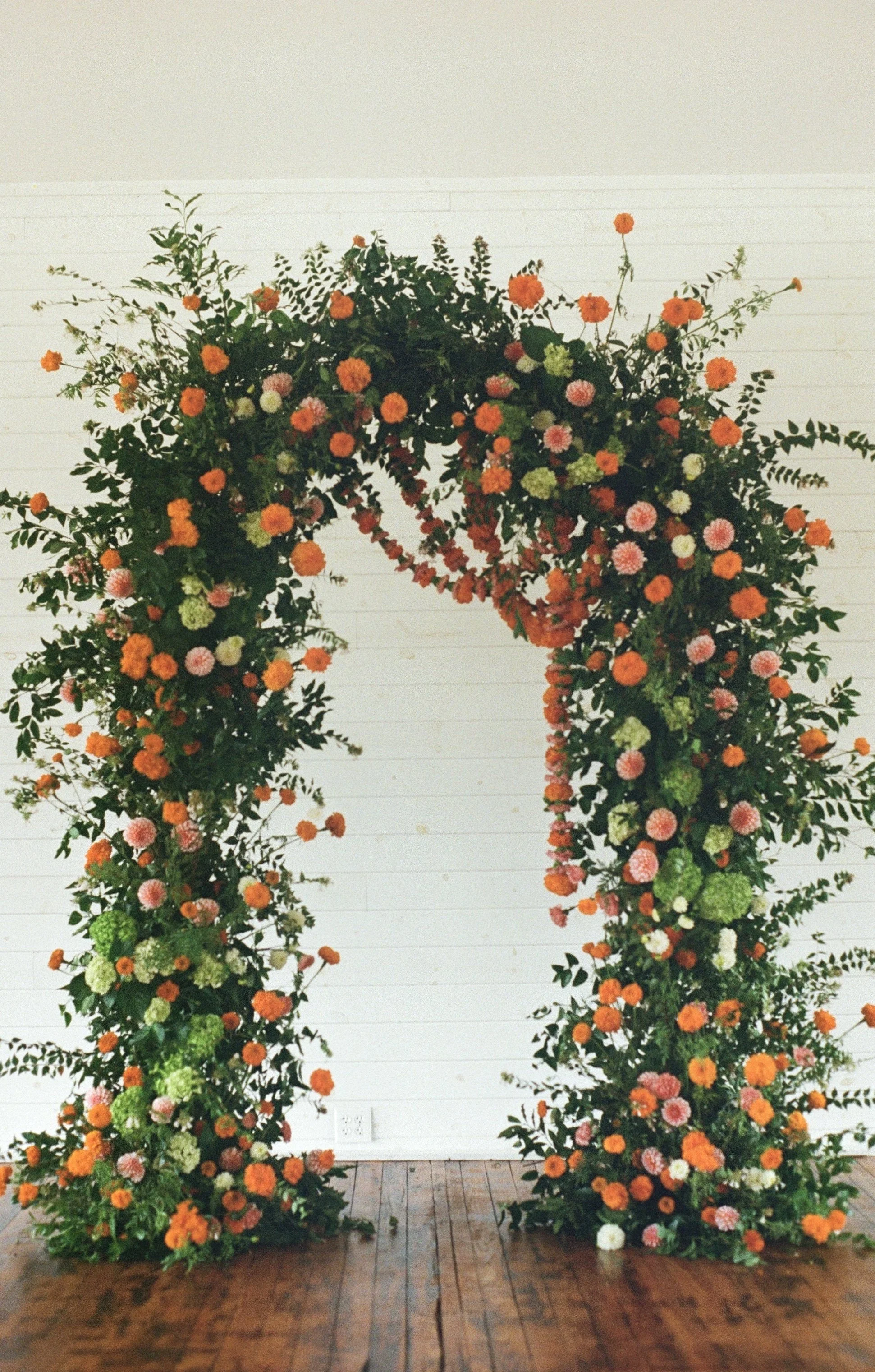A large floral arch with orange, pink, green, and white flowers and green foliage, set against a white wall and wooden floor.