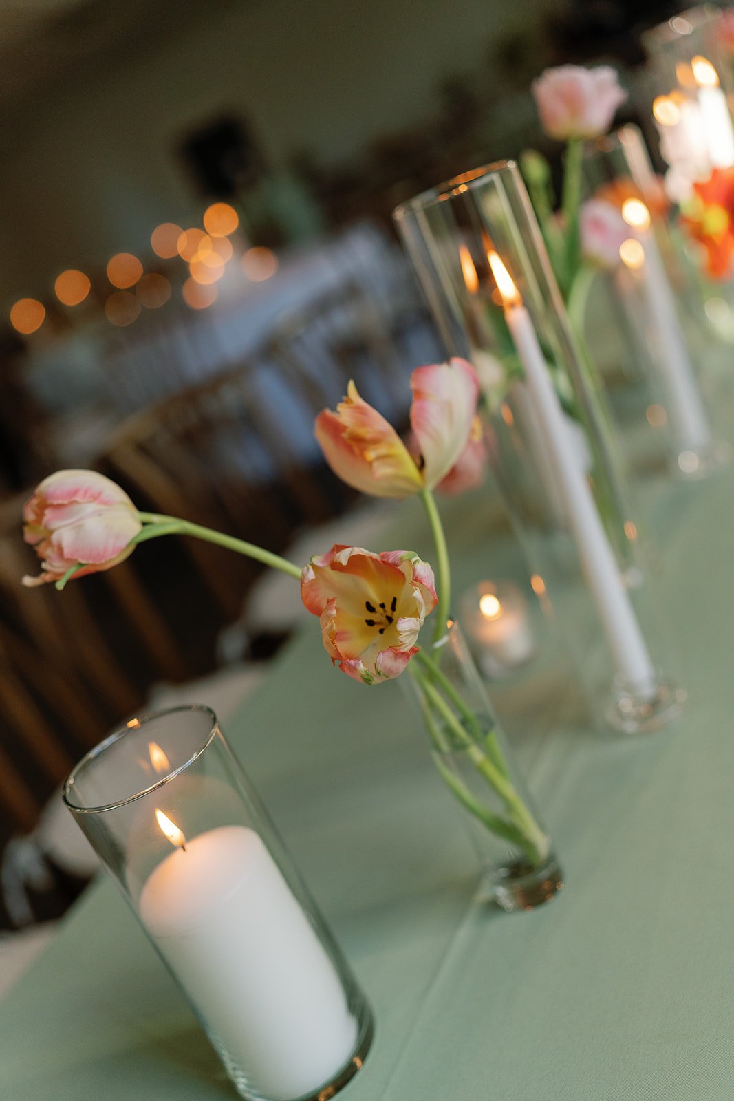 A floral centerpiece with pink and cream tulips in small glass vases, surrounded by tall glass candle holders with lit white candles, on a table decorated for an event or celebration.