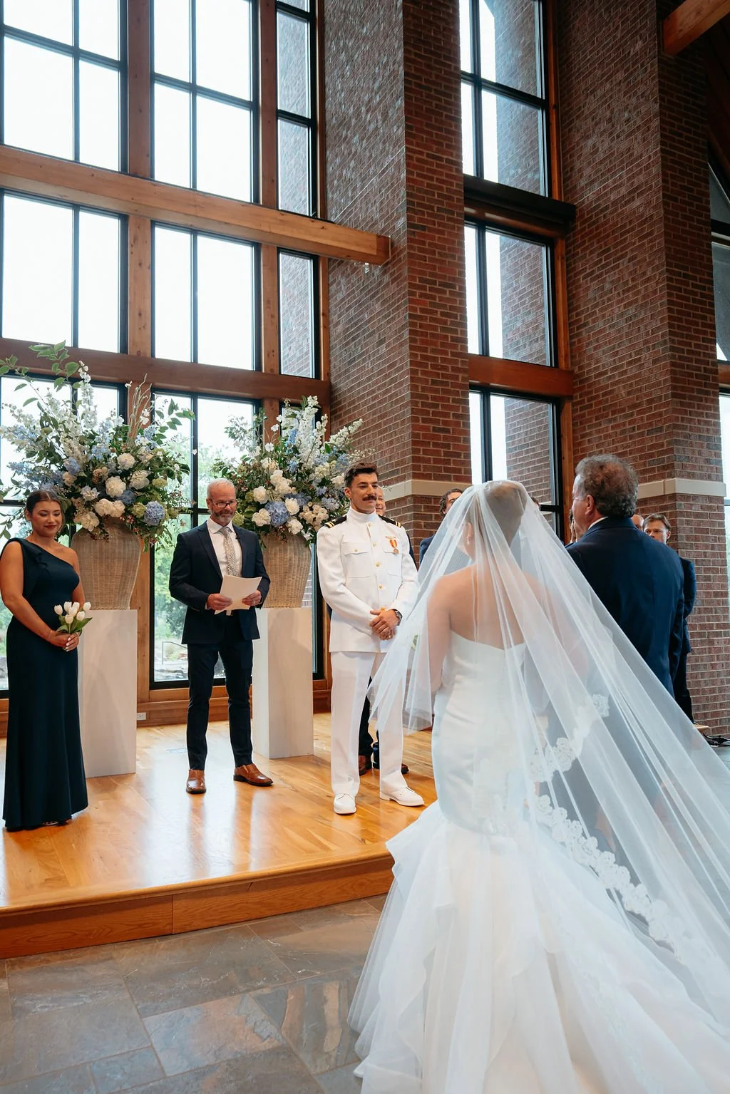 A wedding ceremony taking place indoors in front of large windows with brick and wood framing, featuring a bride and groom facing each other, with the bride wearing a white wedding gown with a long veil, and the groom in a white military uniform. An 