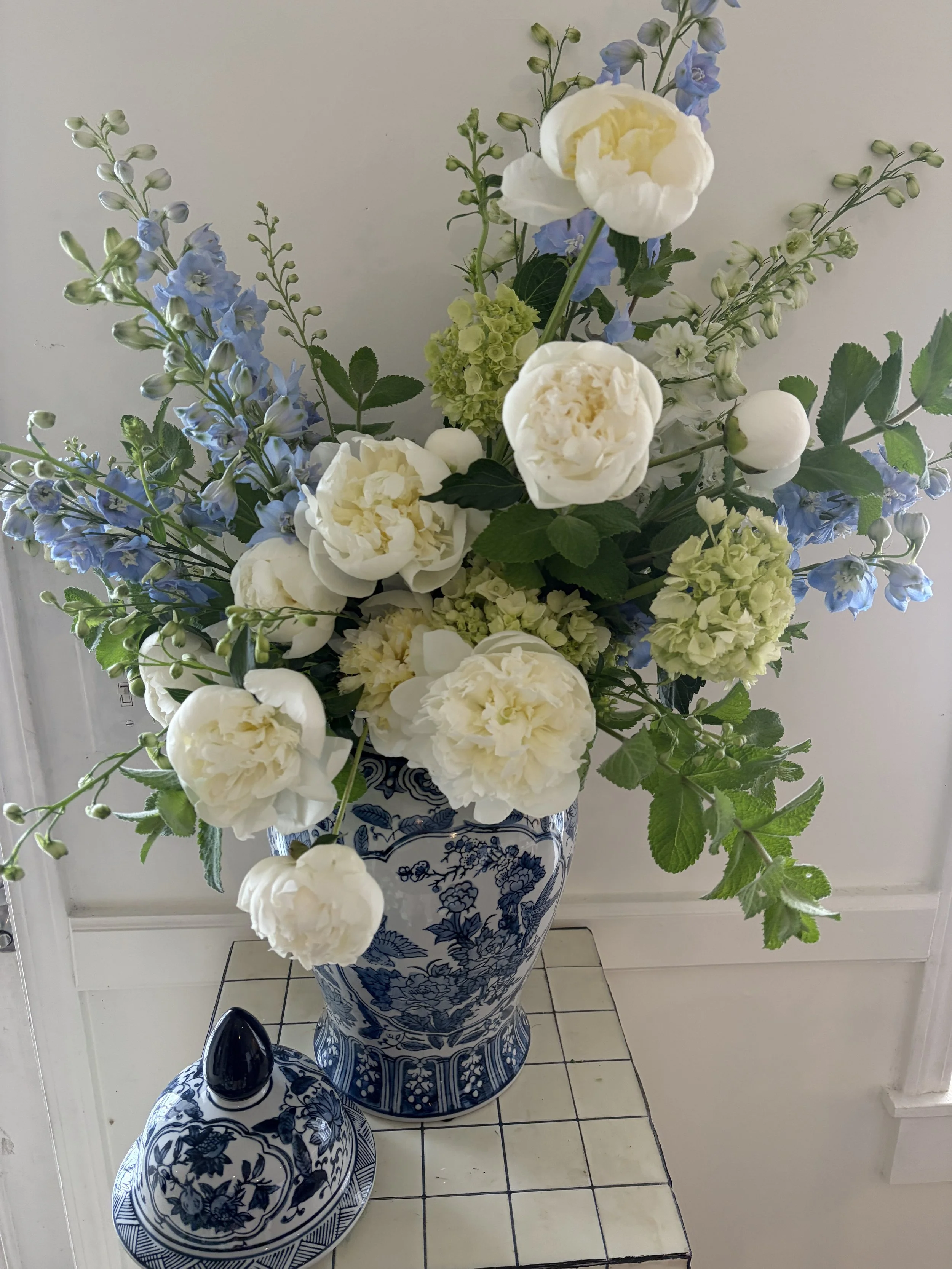 Tall decorative vase with a blue and white floral pattern holding a bouquet of white peonies, blue delphiniums, and other green and white flowers, placed on a tiled floor near a white wall.