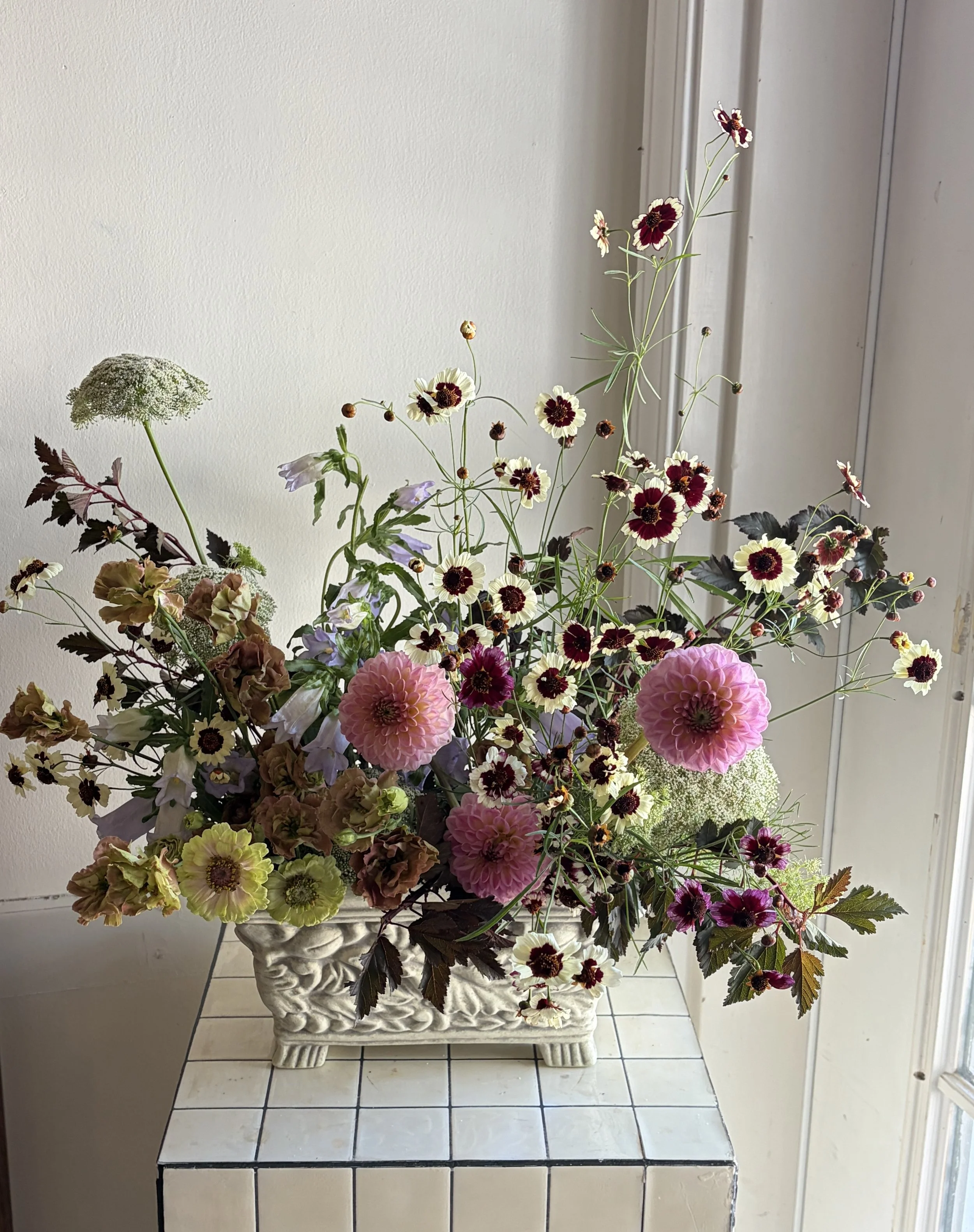 A large floral arrangement in a white ornate ceramic vase, featuring pink dahlias, small white daisies with maroon centers, white Queen Anne's lace, and other colorful flowers, placed on a white tiled surface against a beige wall and near a window.