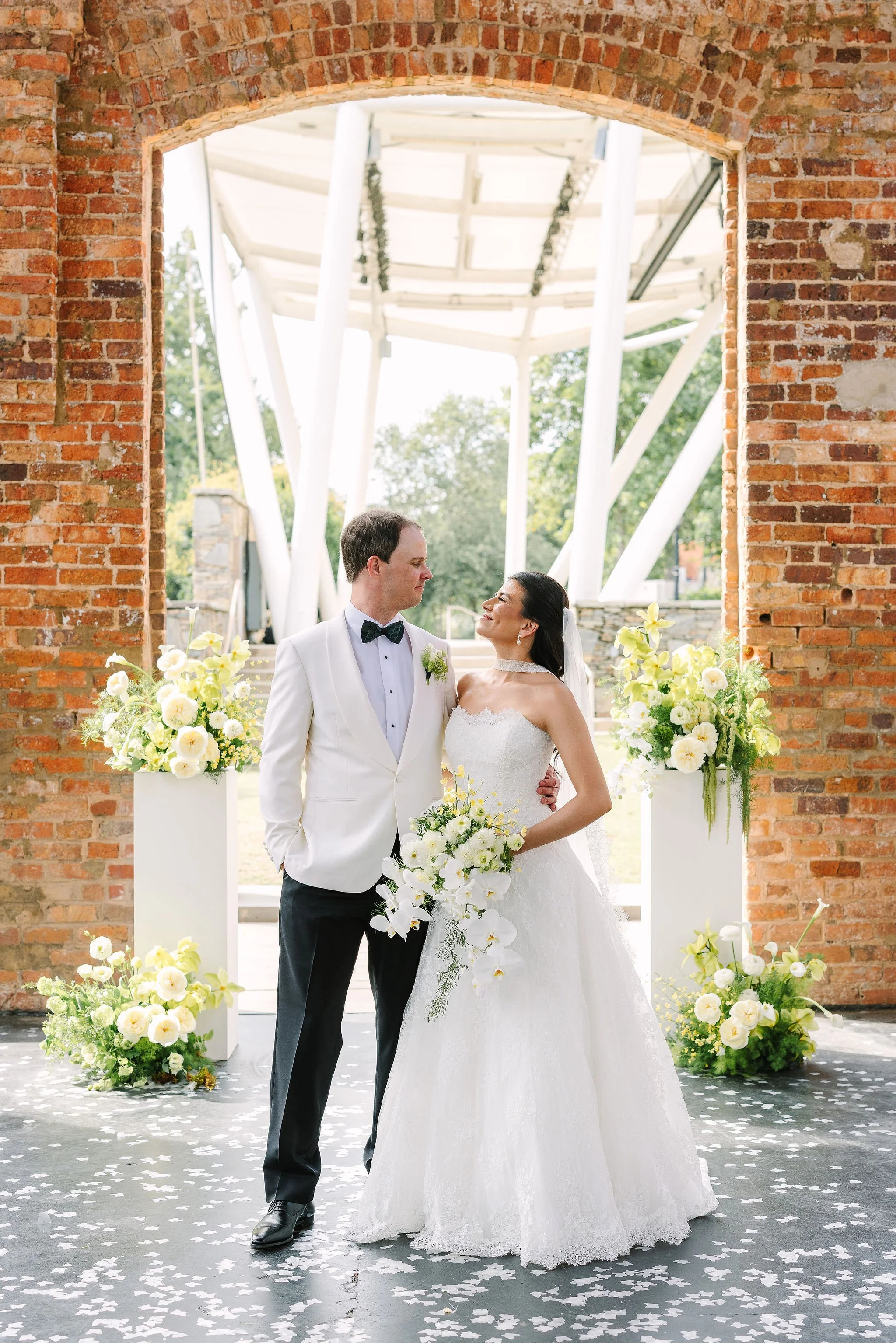 A bride and groom standing together at their wedding ceremony, framed by a brick arch with white and yellow floral arrangements surrounding them. The bride is in a white wedding gown holding a bouquet, and the groom in a white tuxedo with a black bow