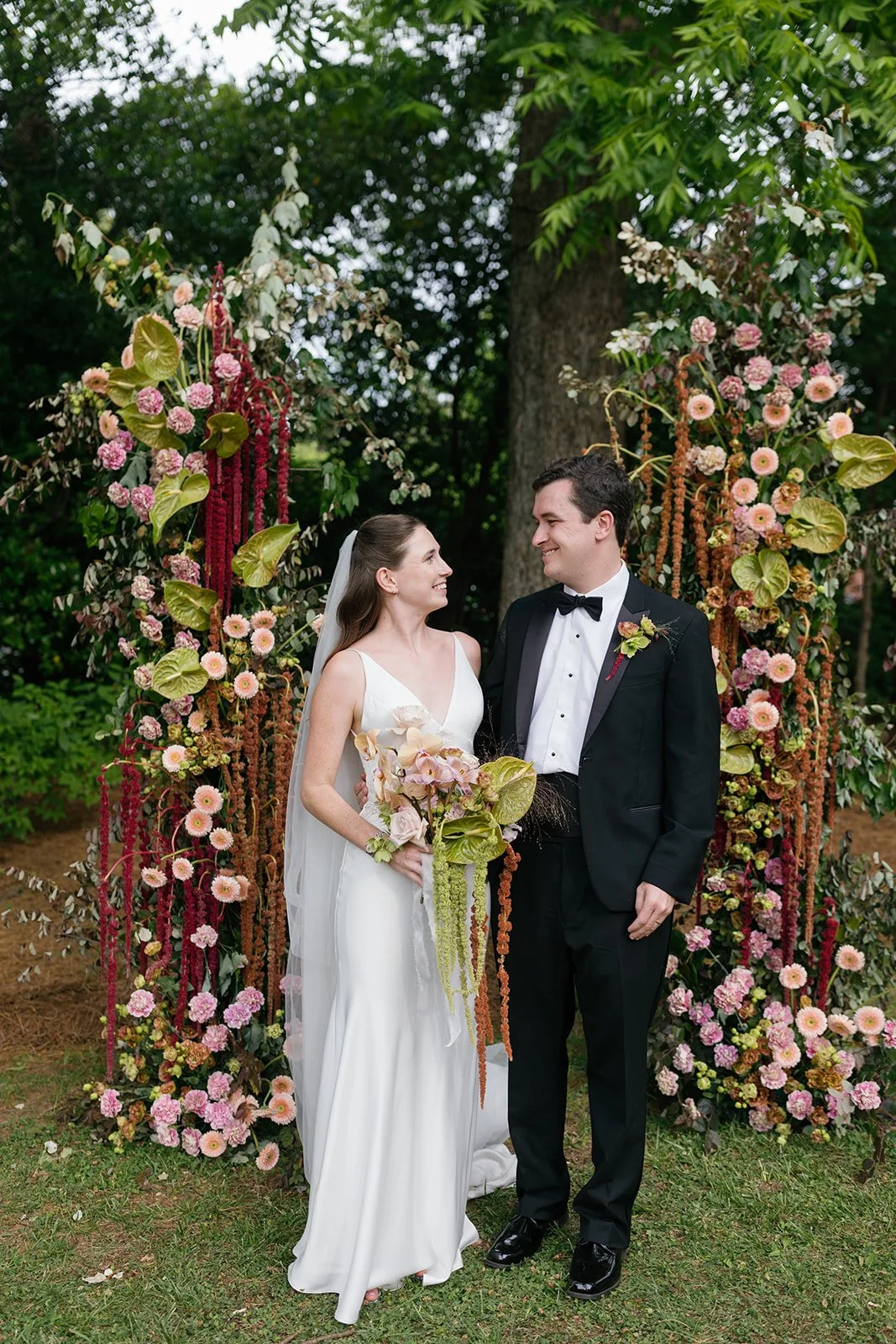 A bride and groom standing together outdoors during a wedding ceremony, surrounded by colorful floral arrangements and greenery, smiling at each other.