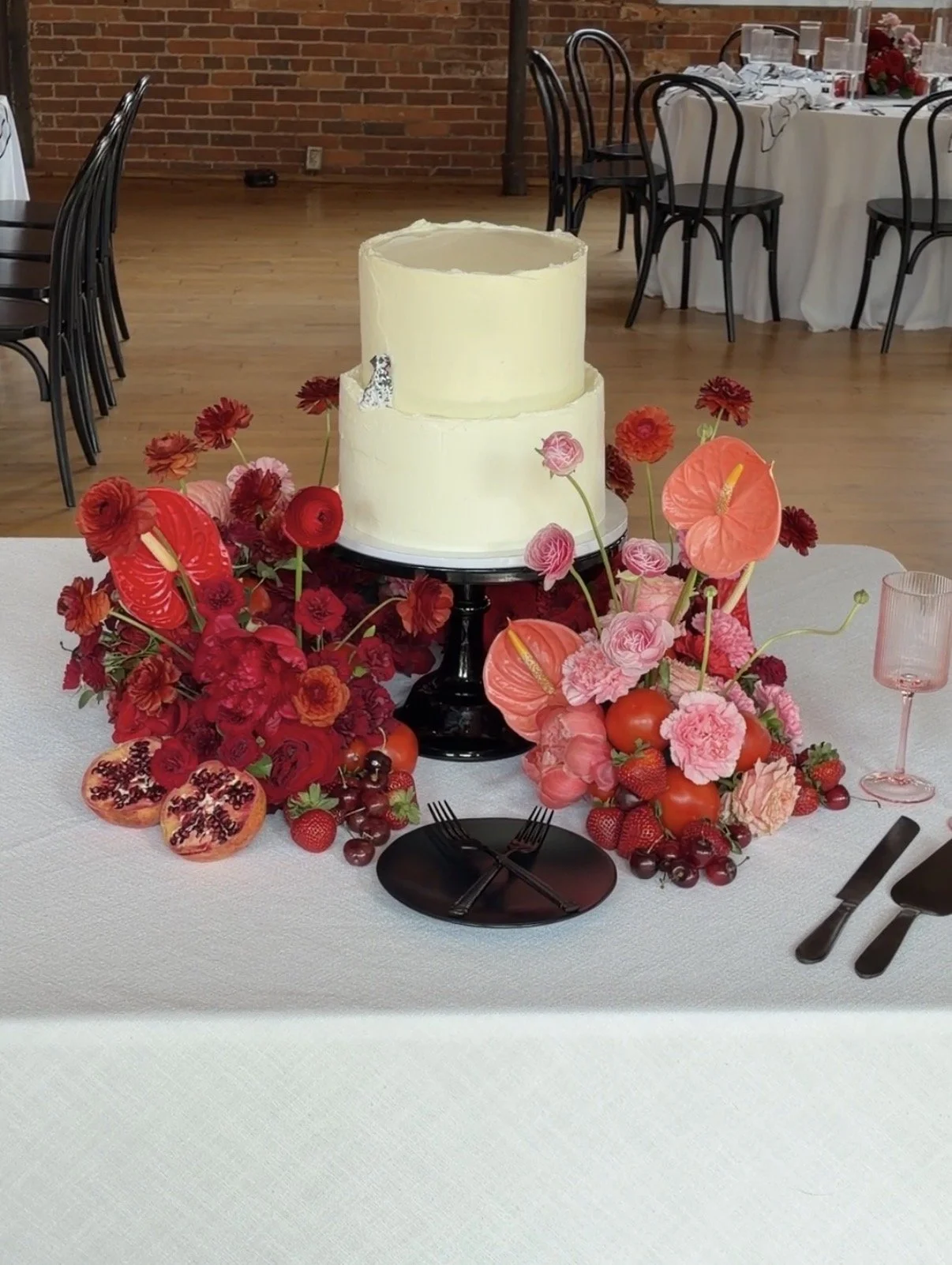 A wedding or celebration table centerpiece with a two-tiered white cake surrounded by red, pink, and orange flowers, strawberries, and pomegranates, set on a white tablecloth with black plates, forks, knives, and a pink glass. In the background are b