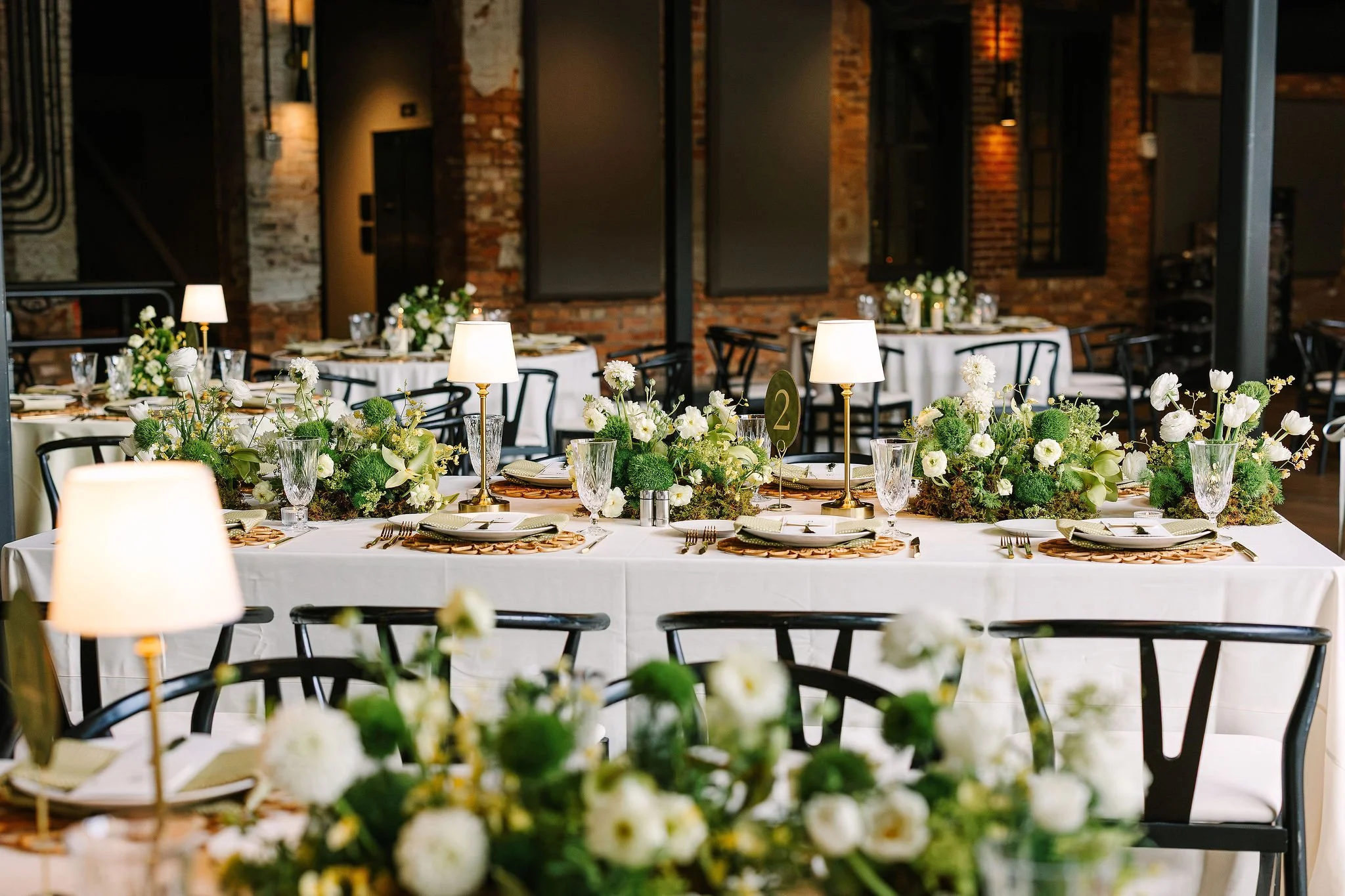 Elegant banquet table decorated with white and green floral centerpieces, surrounded by black chairs, set in an industrial-style venue with exposed brick walls and warm lighting.