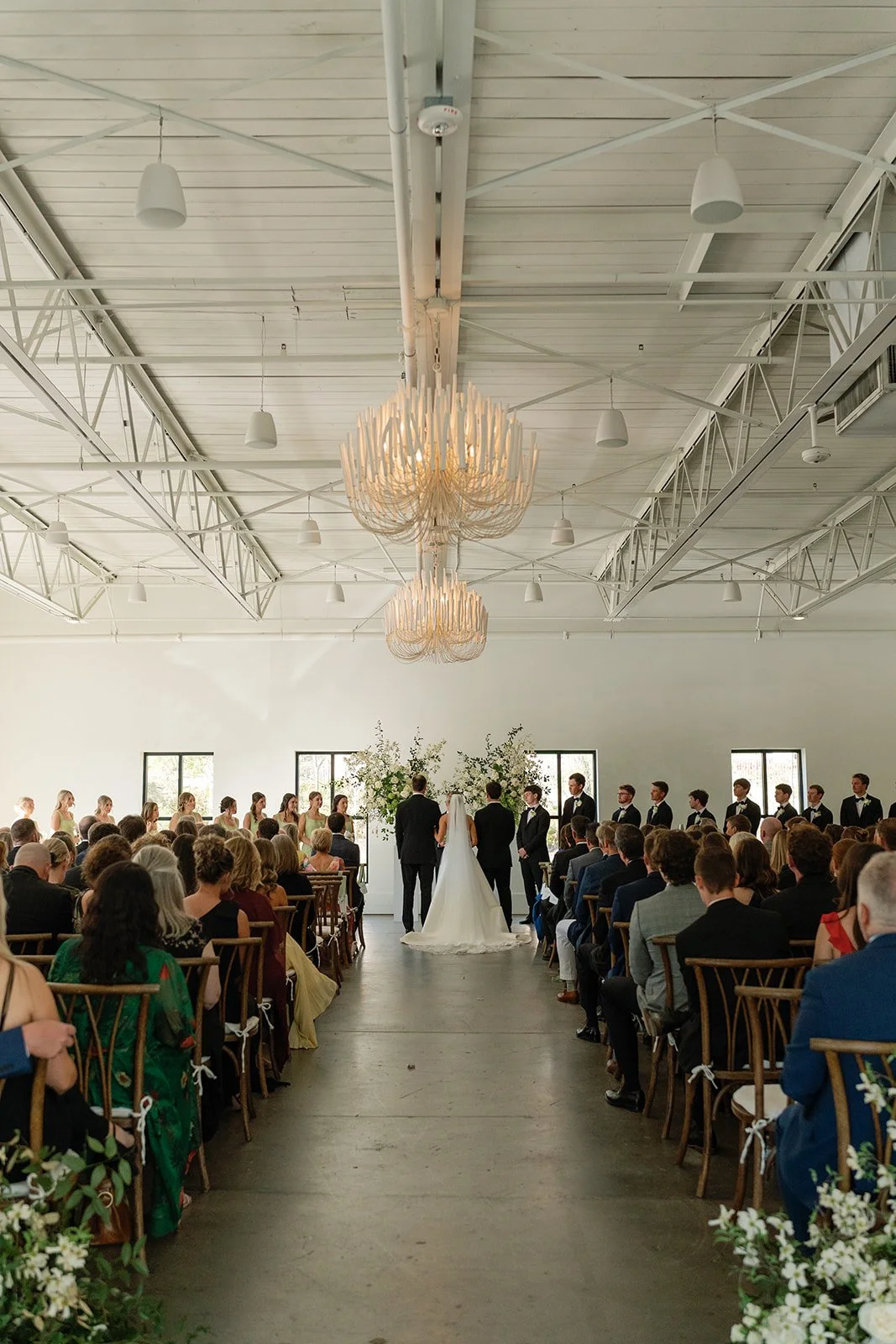 Indoor wedding ceremony with bride, groom, and officiant at the altar, surrounded by wedding party and seated guests, large chandelier, and white walls.