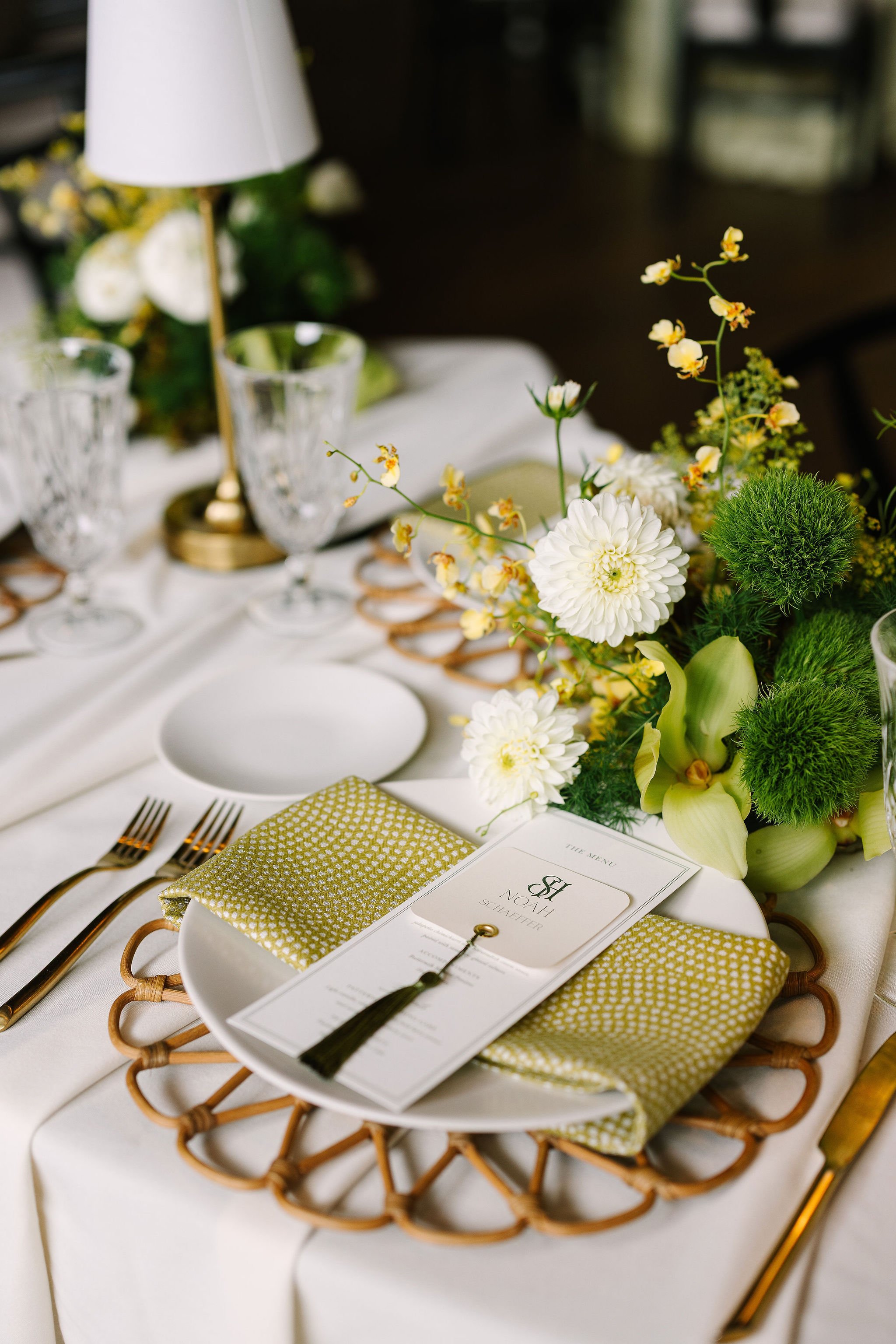 A table setting with white plates, gold utensils, a menu card, a gold patterned napkin, and a floral centerpiece featuring white flowers and green foliage. The tablecloth is white.