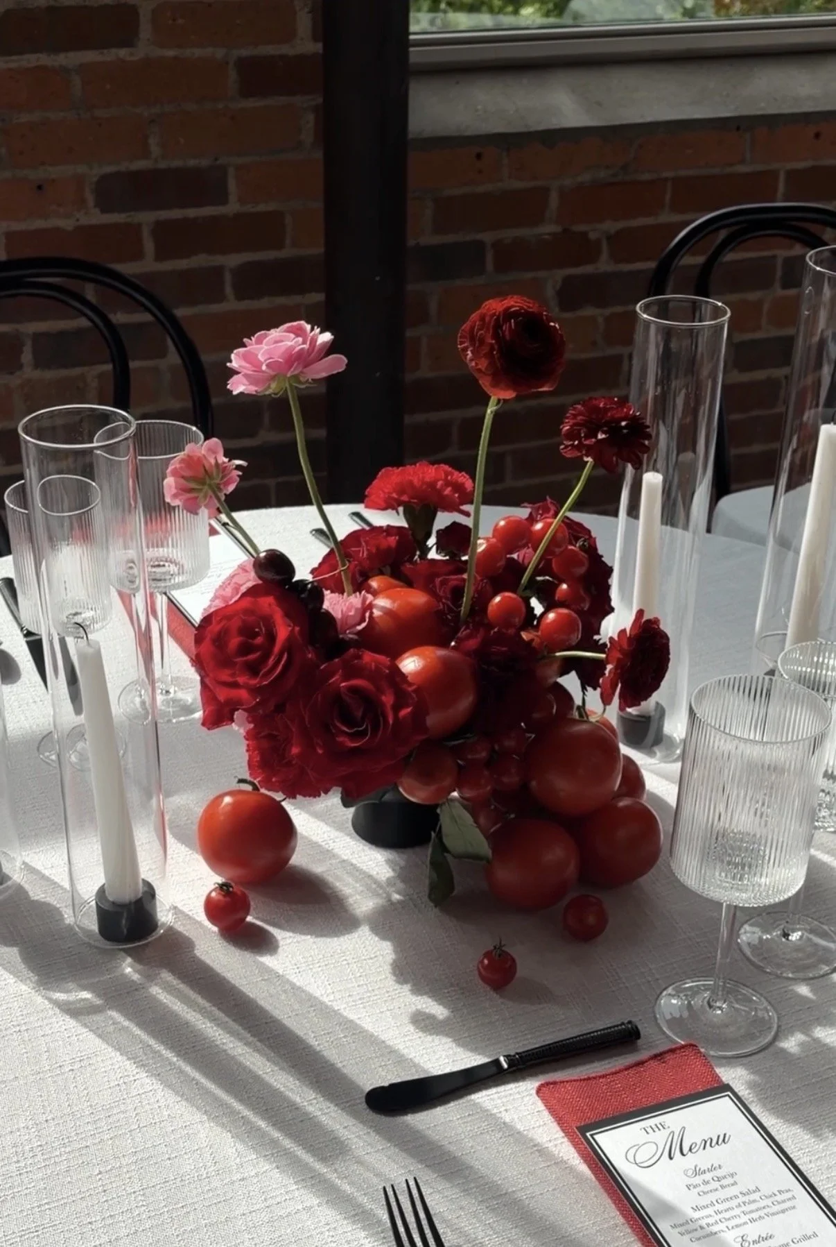 A table decorated with a bouquet of red and pink flowers and cherry tomatoes, surrounded by candle holders and glassware, set for a meal with a printed menu.