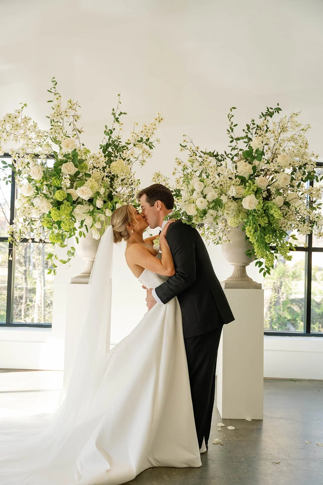 A bride and groom kiss in front of large floral arrangements in a bright, modern wedding venue.