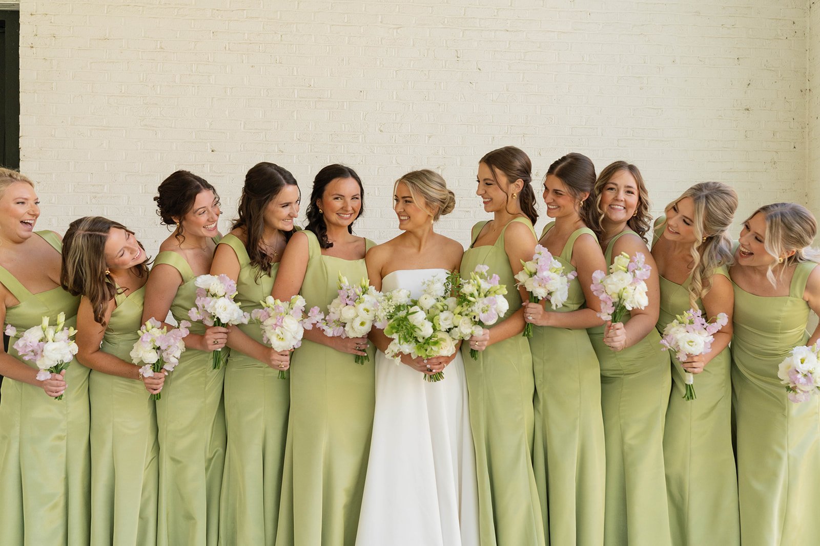 A bride and her bridesmaids in matching light green dresses holding bouquets, standing against a white brick wall, smiling at each other at a wedding.