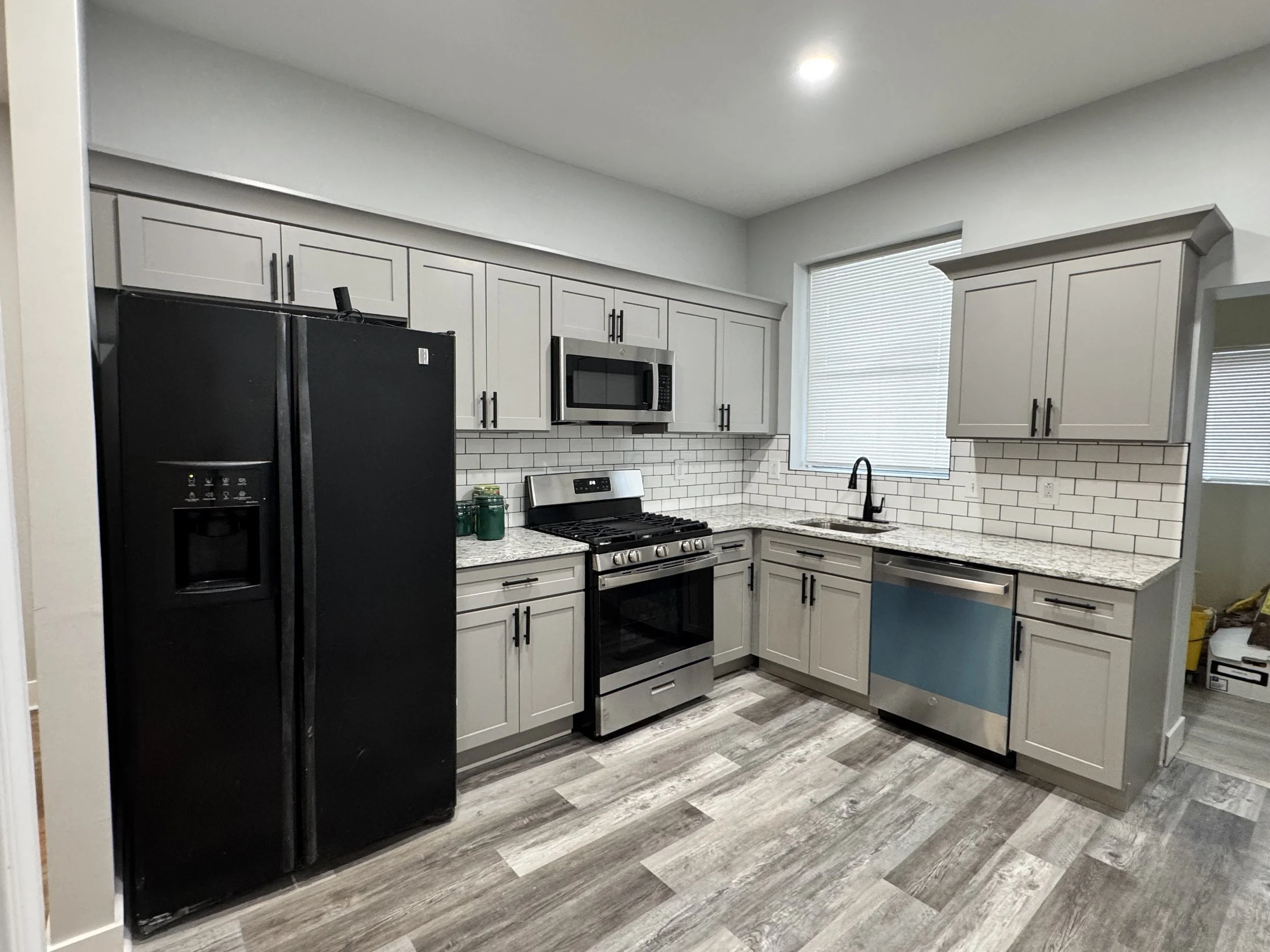 Modern kitchen with black refrigerator, stainless steel stove and microwave, gray cabinets, white subway tile backsplash, granite countertops, and wood-look flooring.