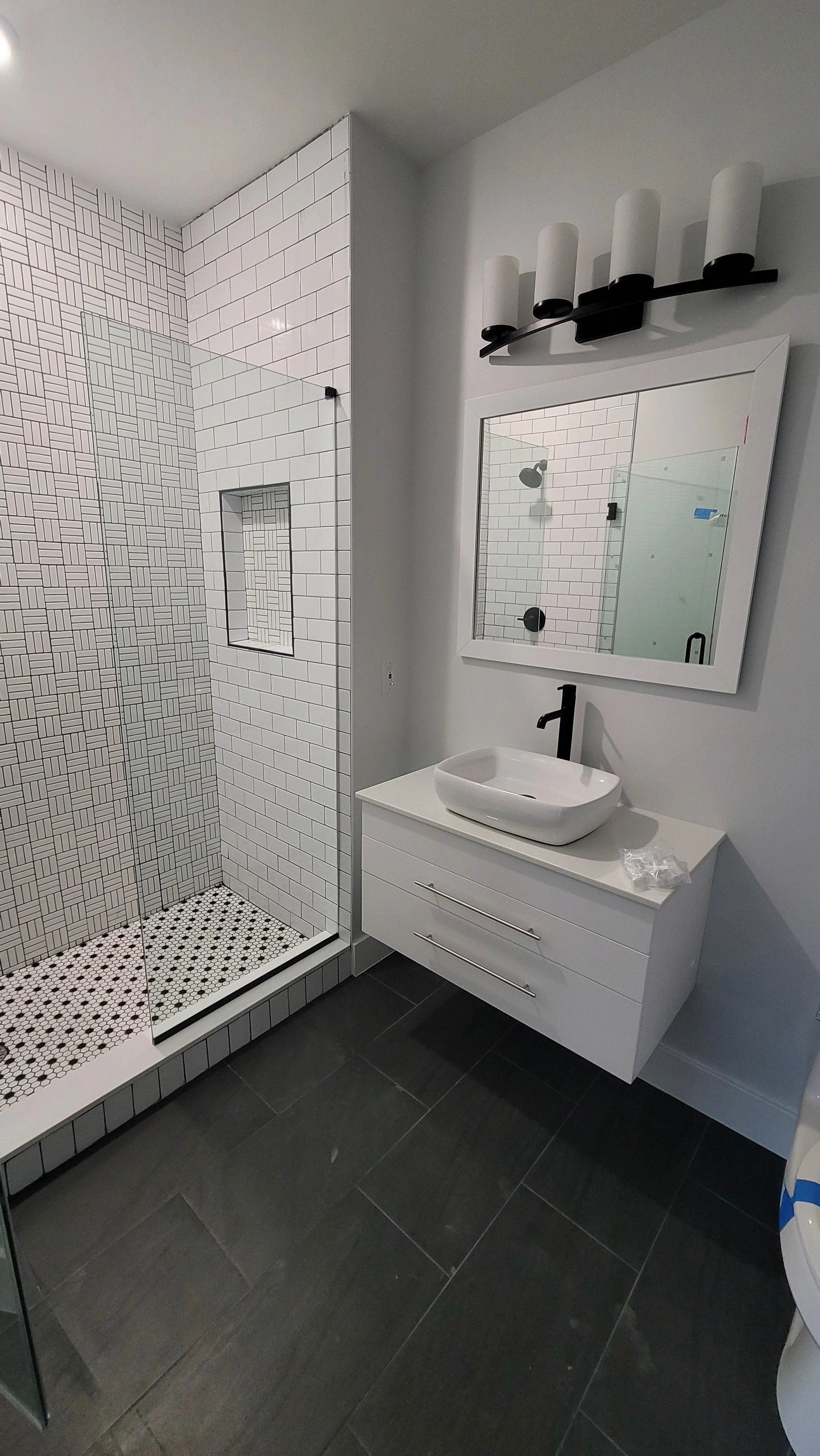 Modern bathroom with white vanity sink, black faucet, large mirror, style lighting fixture, and walk-in shower with black and white tiles.