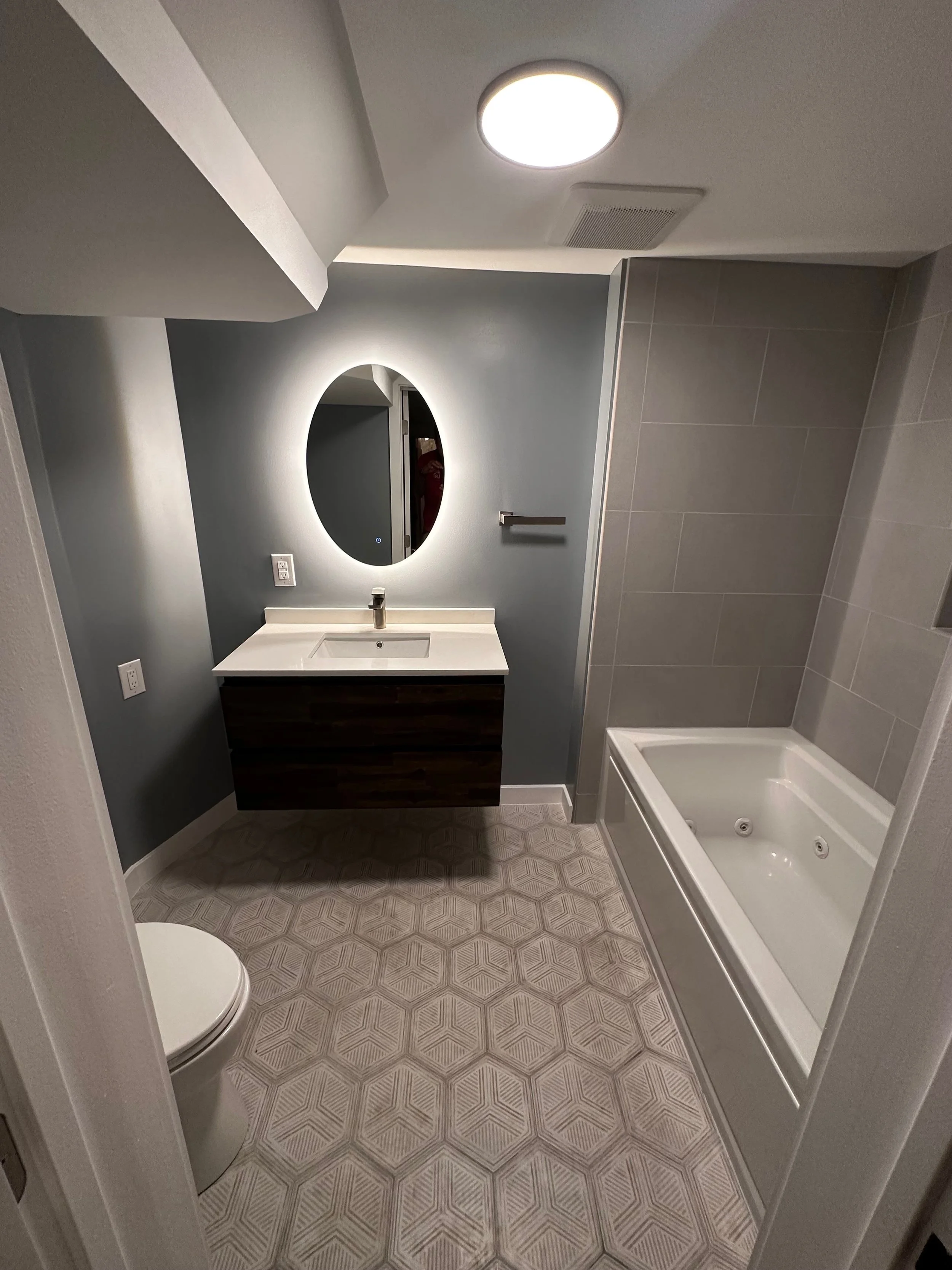 Modern bathroom featuring a vanity with a mirror, a toilet, and a bathtub with jets, with gray and white tiles and wall-mounted fixtures.