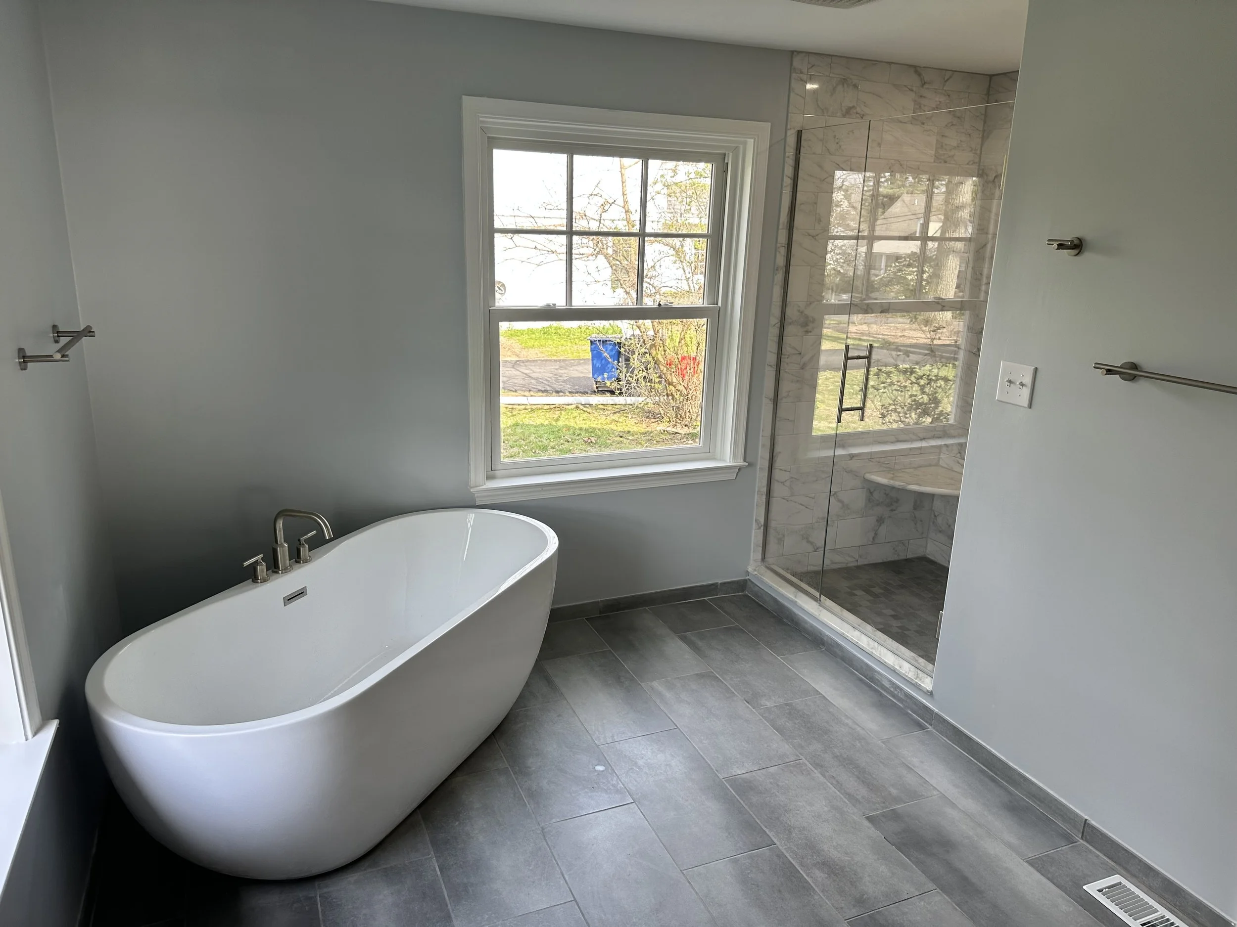 Bathroom with a standalone bathtub, a window overlooking the outside, and a glass-enclosed shower with marble tiles.